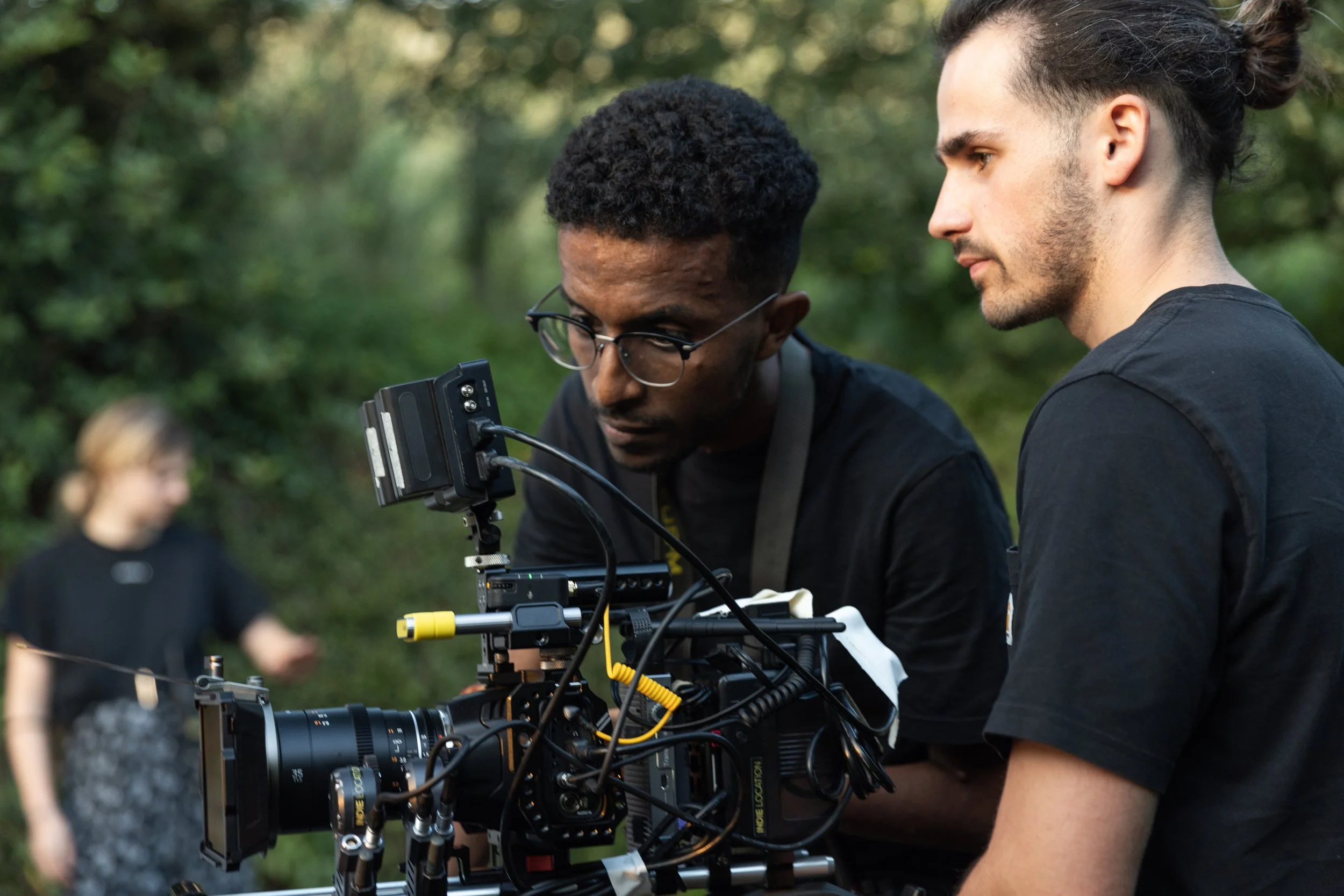 Deux hommes en noir travaillent avec une caméra professionnelle en plein air, avec une femme floue en arrière-plan.