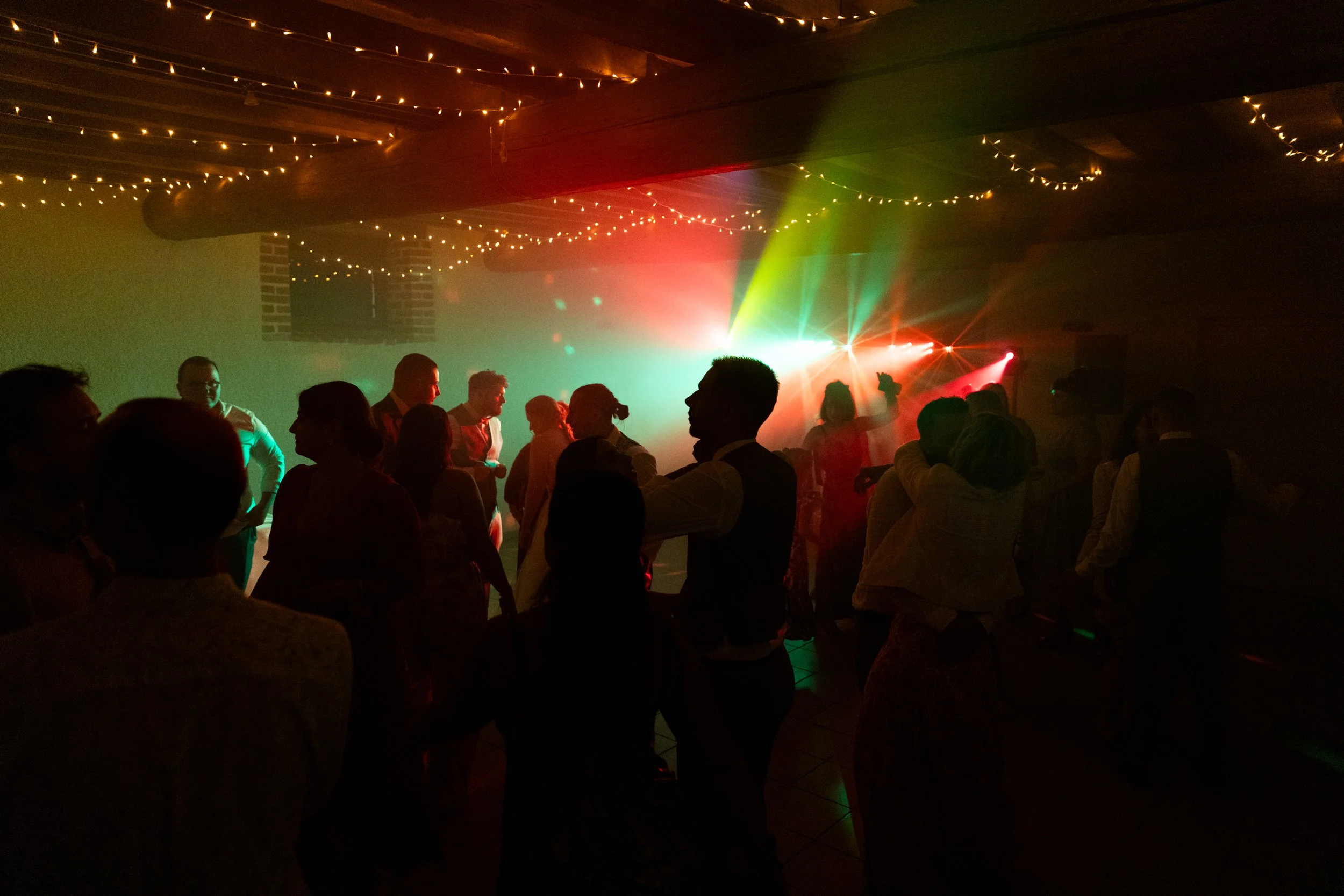 Groupe de personnes dansant dans une salle de fête avec des lumières colorées et guirlandes lumineuses au plafond.