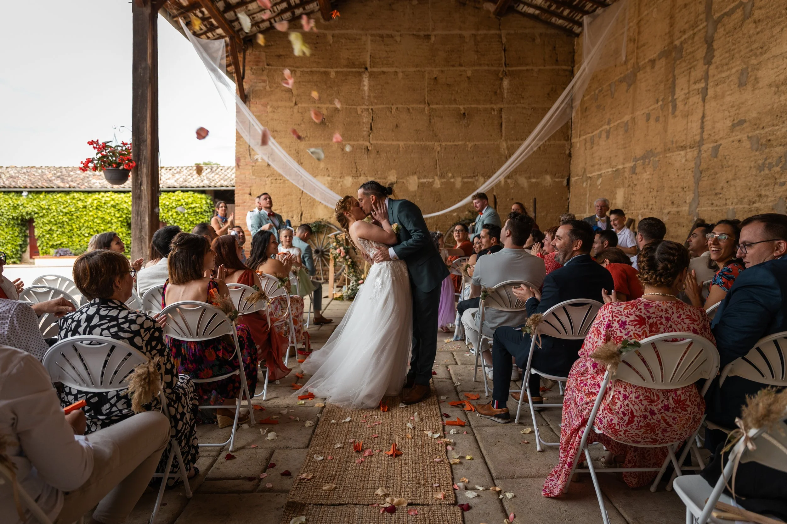 Un mariage avec la mariée et le marié s'embrassant au centre d'une cérémonie en plein air, entourés d'invités assis, dans un lieu rustique avec des murs en pierre et des décorations florales.