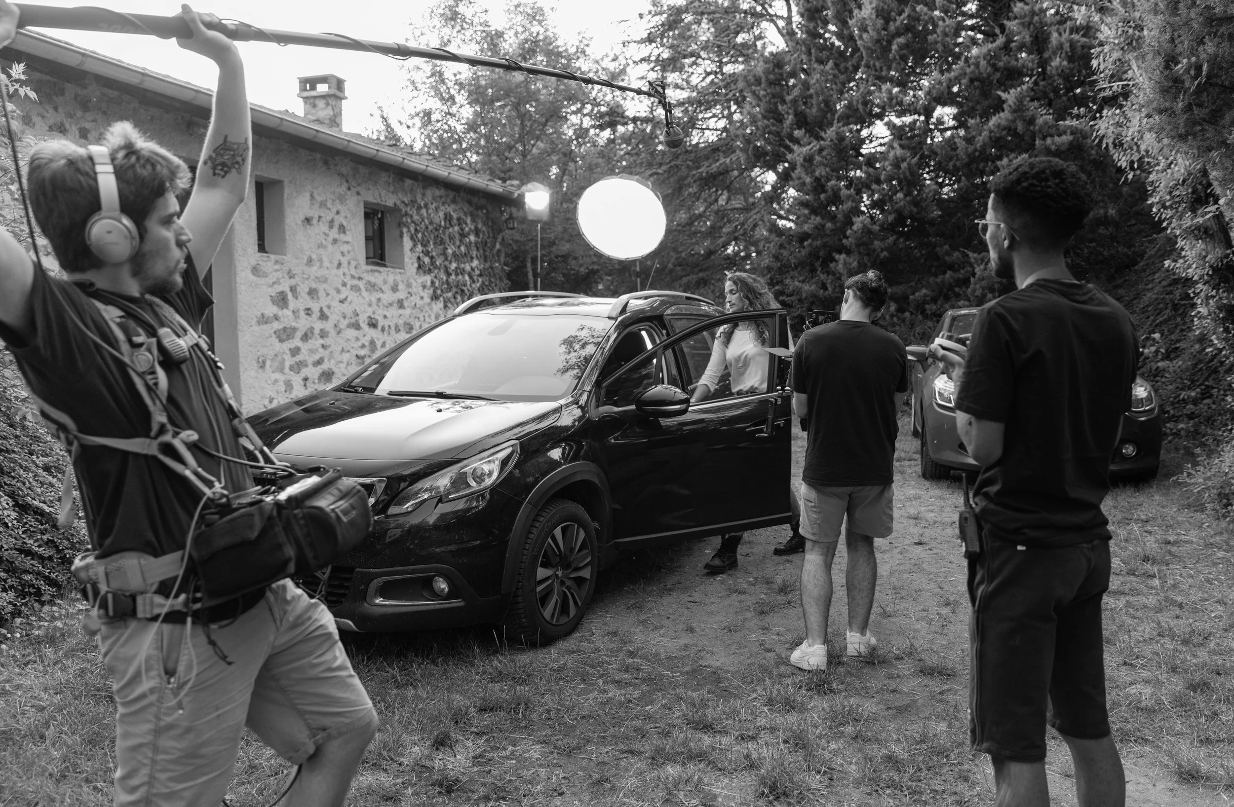 Une équipe de tournage filme une scène à l'extérieur avec une voiture noire. Deux hommes, une femme et un technicien avec du matériel de tournage sont présents.