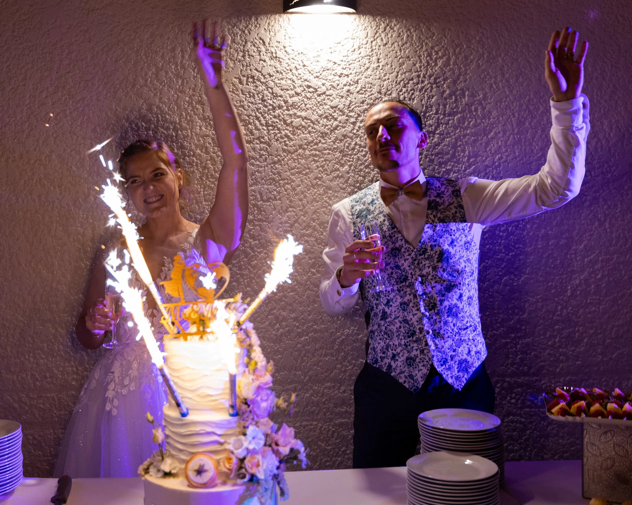 Un couple lors d'un mariage, la femme porte une robe de mariée blanche et le marié porte un gilet à motifs et une veste, ils tiennent des verres et la femme change d'expressions avec un gâteau de mariage en premier plan, des fusées éclairantes sur le