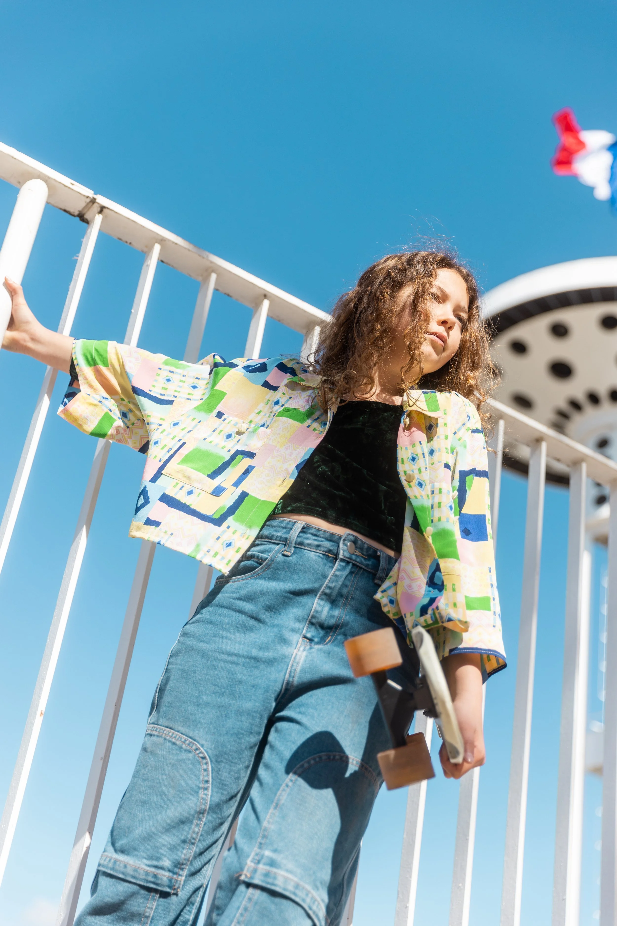 Une jeune fille avec des cheveux bouclés, vêtue d'une chemise colorée, d'un pantalon en denim et tenant un skateboard, se tient près d'une clôture, avec une grande roue visible en arrière-plan contre un ciel bleu.
