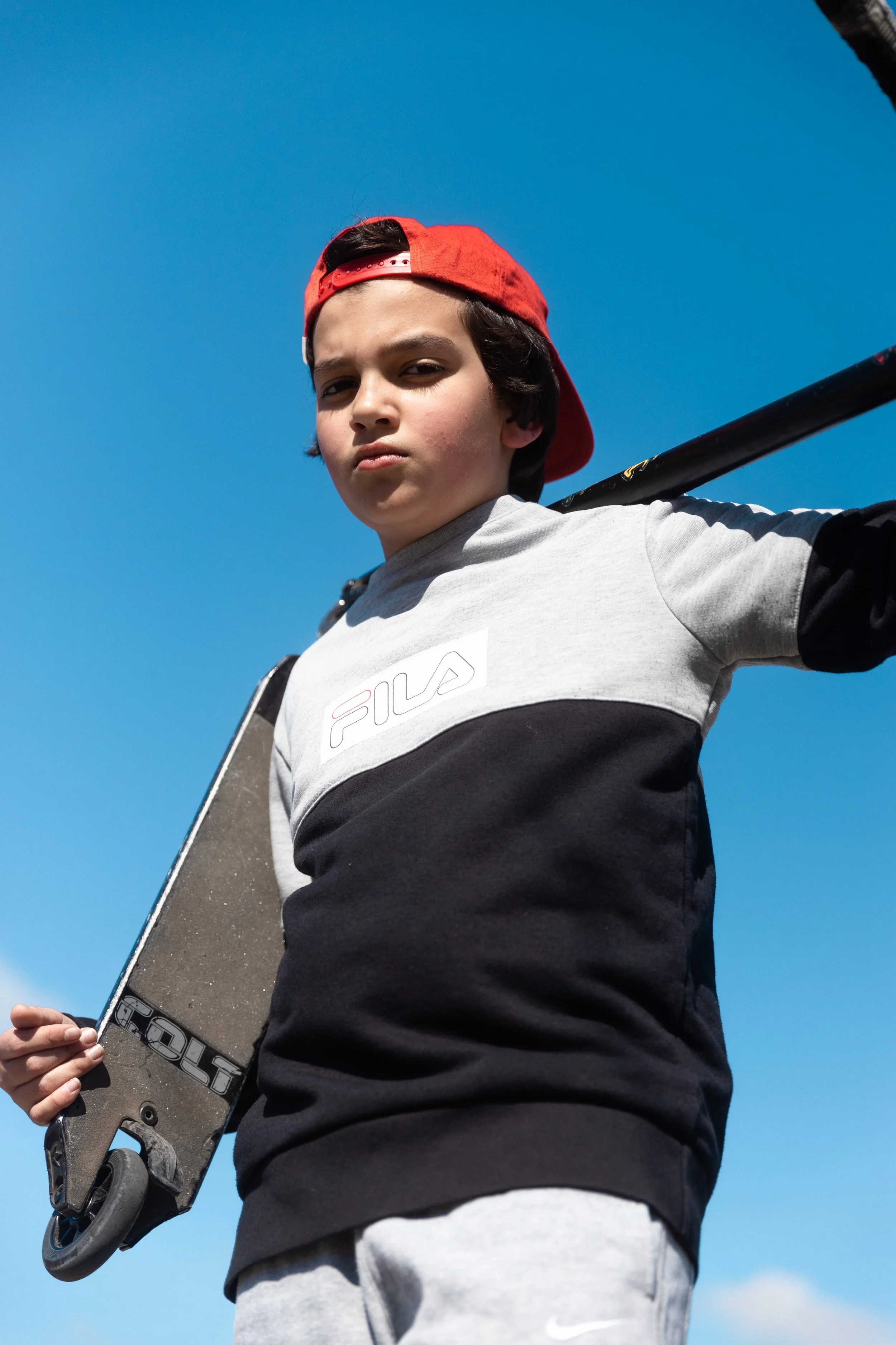 Un garçon portant une casquette rouge à l'envers, tenant un skateboard, regardant vers l'avant, avec un ciel bleu clair en arrière-plan.