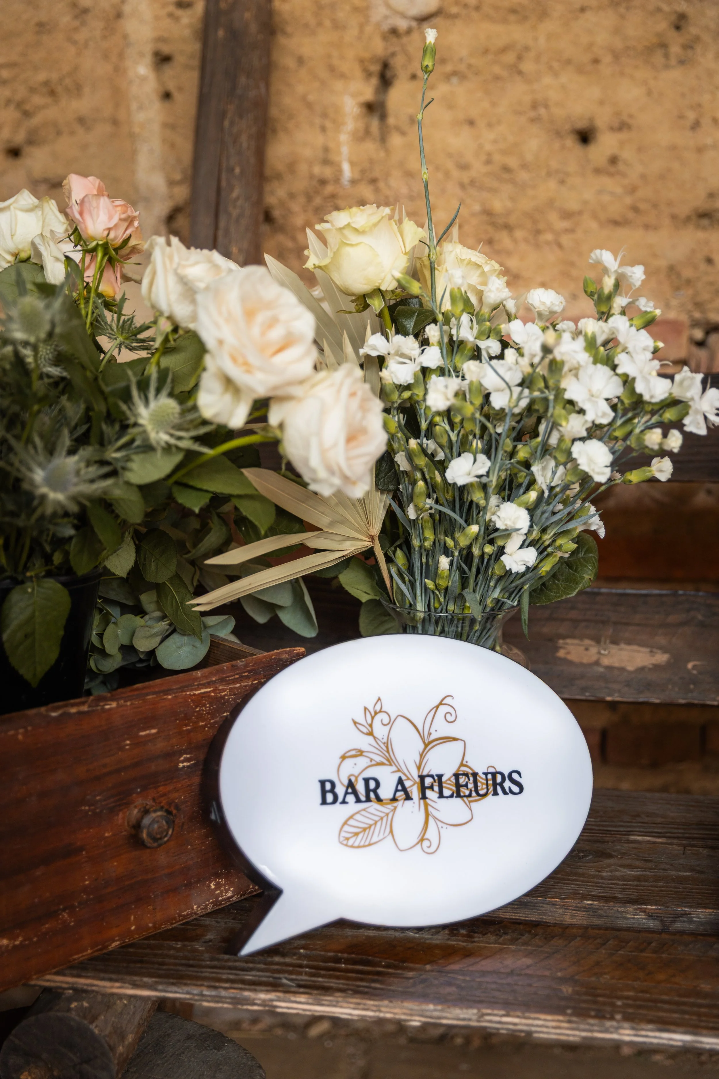 Un arrangement floral composé de roses blanches, de petites fleurs blanches et de feuillage, avec une plaque en céramique indiquant 'BARA FLEURS'.