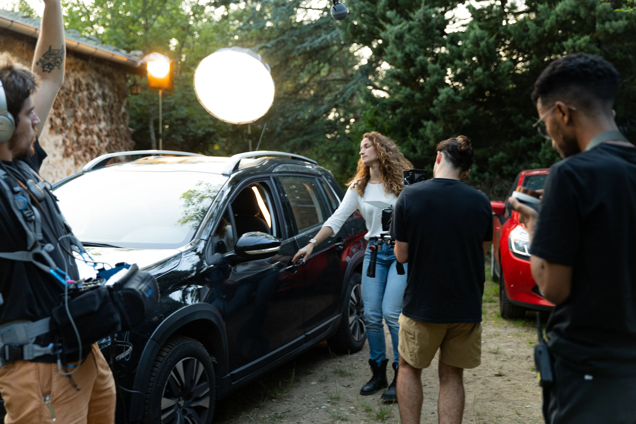 Une équipe de tournage filme une femme qui ouvre la portière d'une voiture noire, avec un fond de nature et un éclairage professionnel.