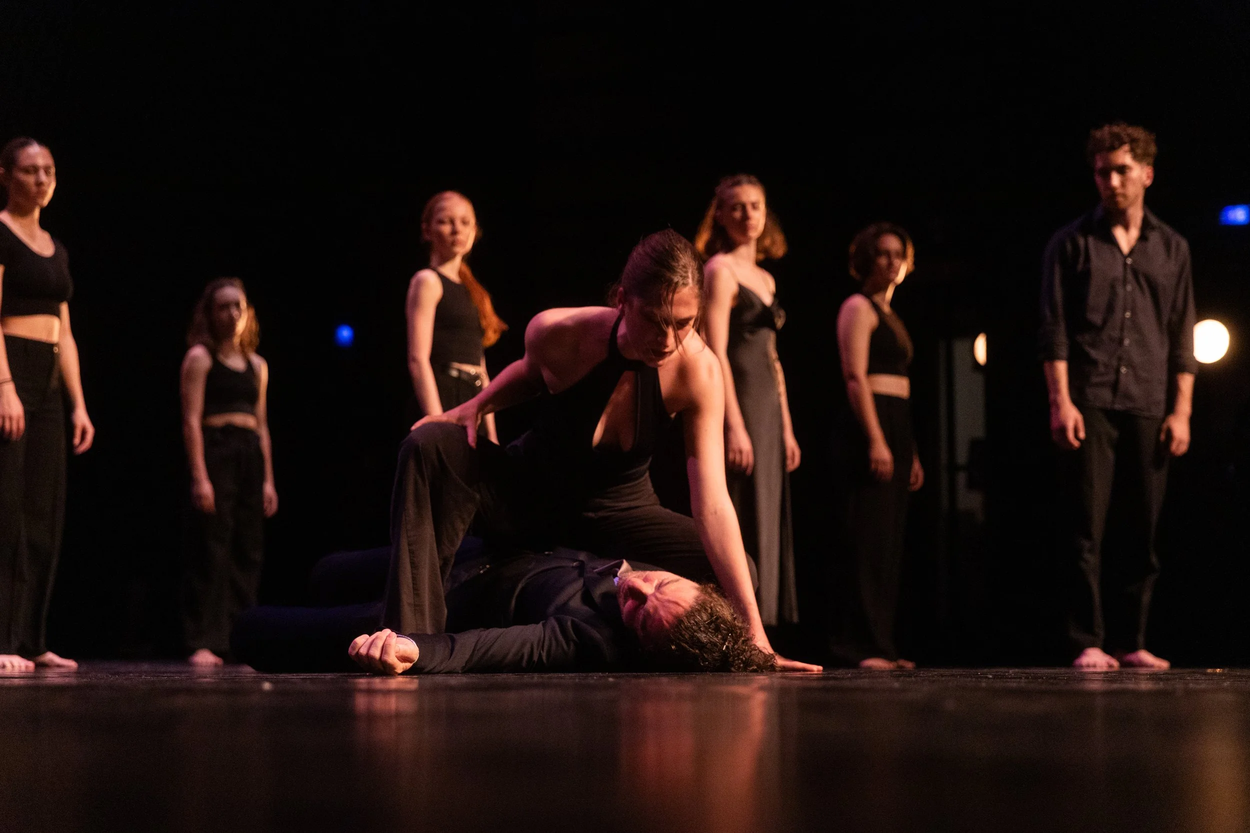 Une scène de danse contemporaine avec plusieurs danseurs en noir, où une femme est en position dominante sur un homme allongé au sol, dans une pièce sombre avec un éclairage de scène.