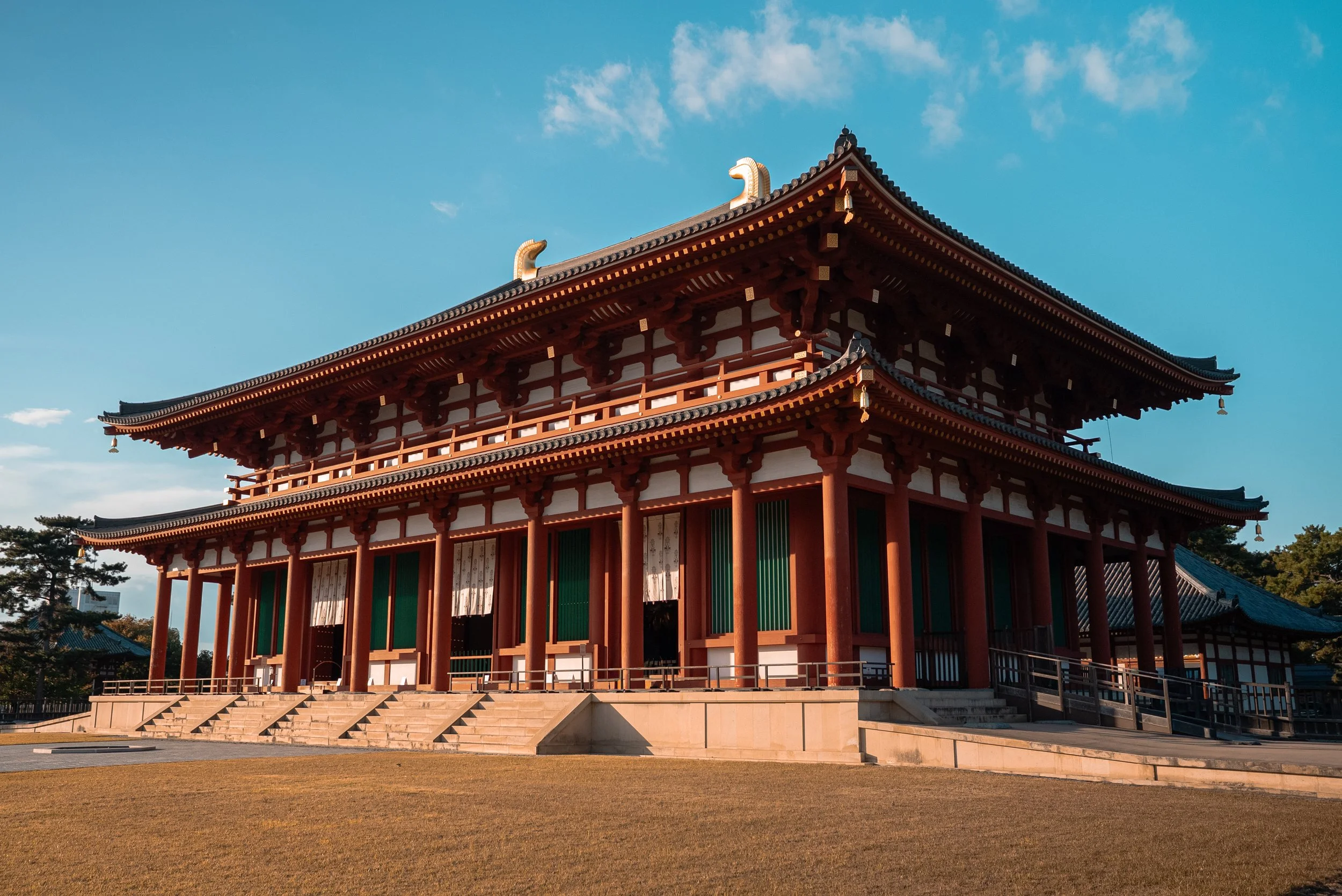 Un bâtiment traditionnel japonais en bois rouge avec un toit en tuiles, entouré d'un jardin et sous un ciel bleu clair.