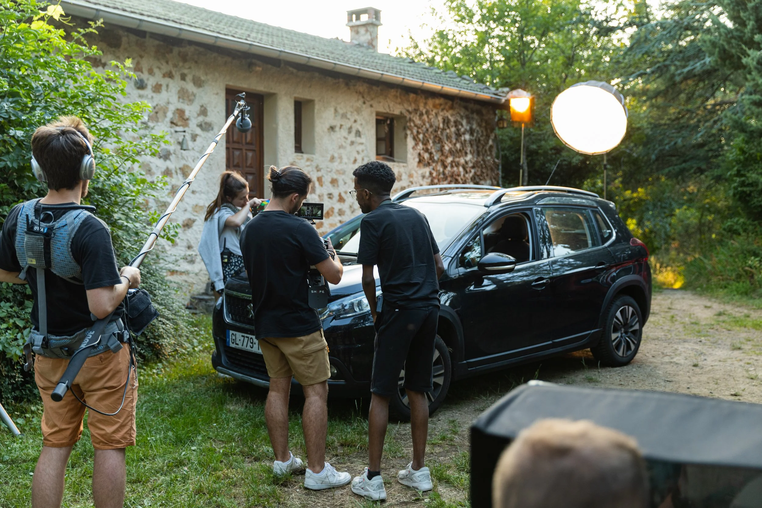 Une équipe de tournage réalise une scène devant une voiture noire garée devant une maison en pierre, avec un éclairage professionnel et du matériel de tournage dans un environnement extérieur verdoyant.