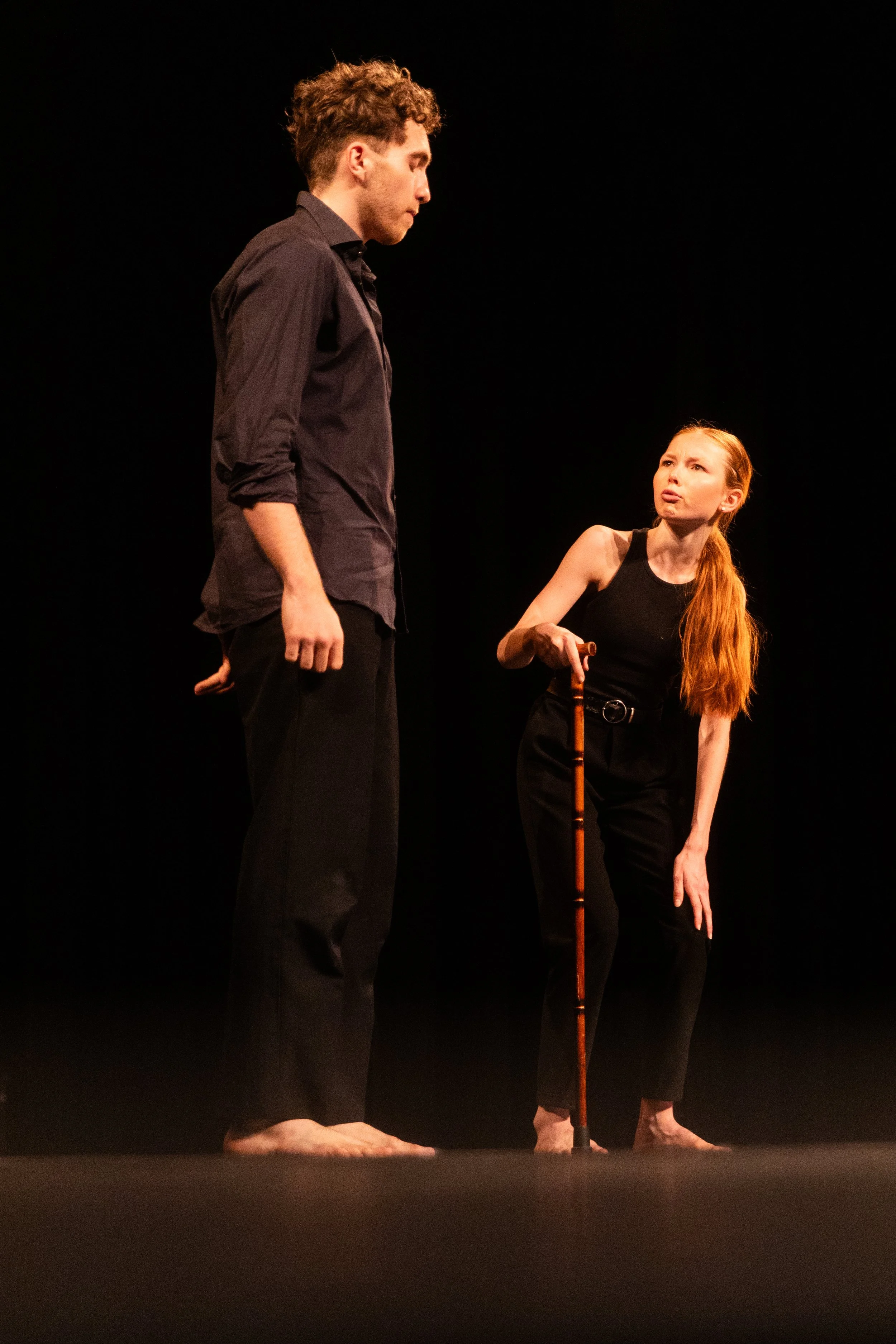 Deux jeunes acteurs sur scène, l'un homme et une femme avec une canne, en pleine performance théâtrale, dans une ambiance sombre.