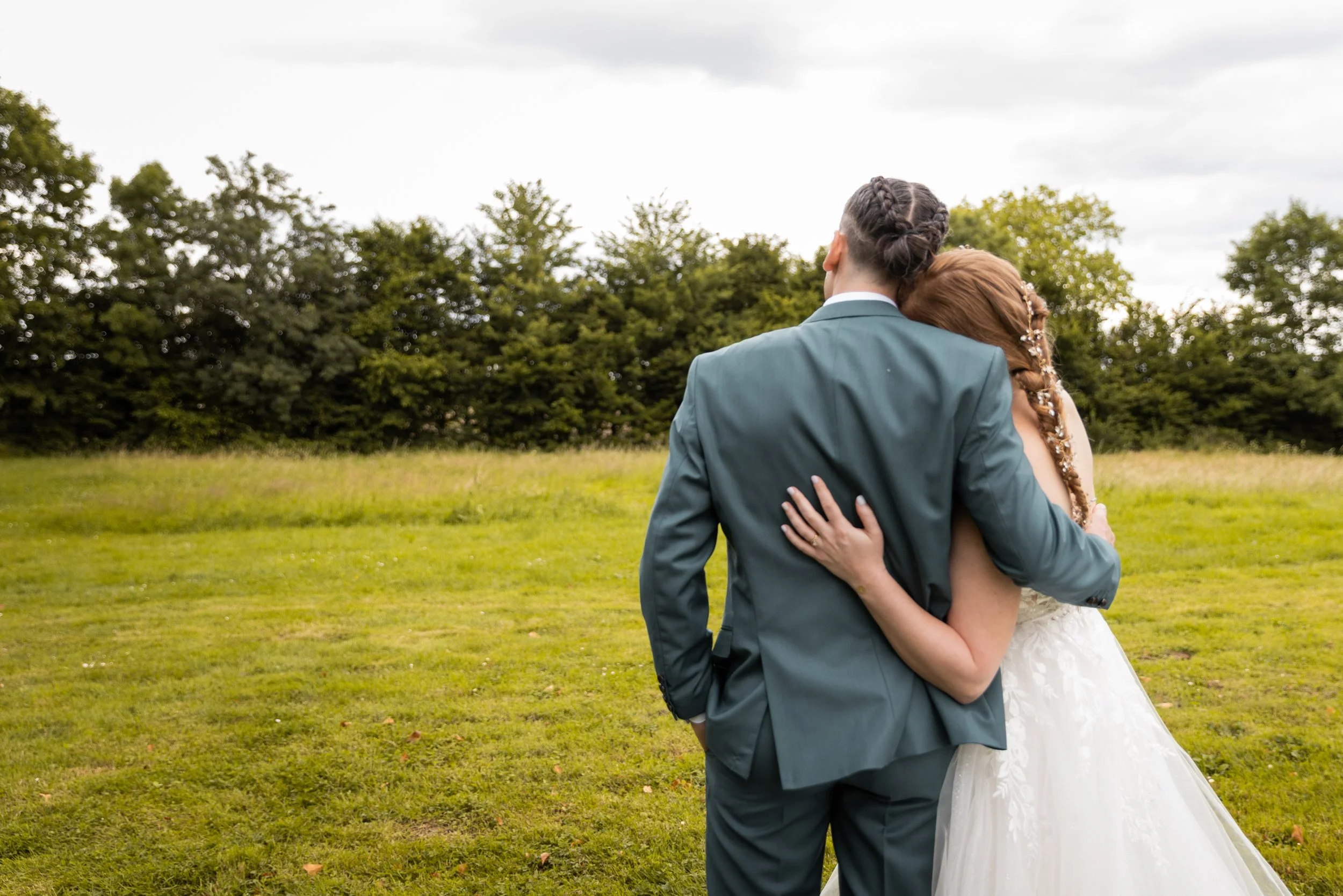 Un couple en mariage, vue de dos, embrassant dans un champ vert avec des arbres au fond, ciel nuageux.