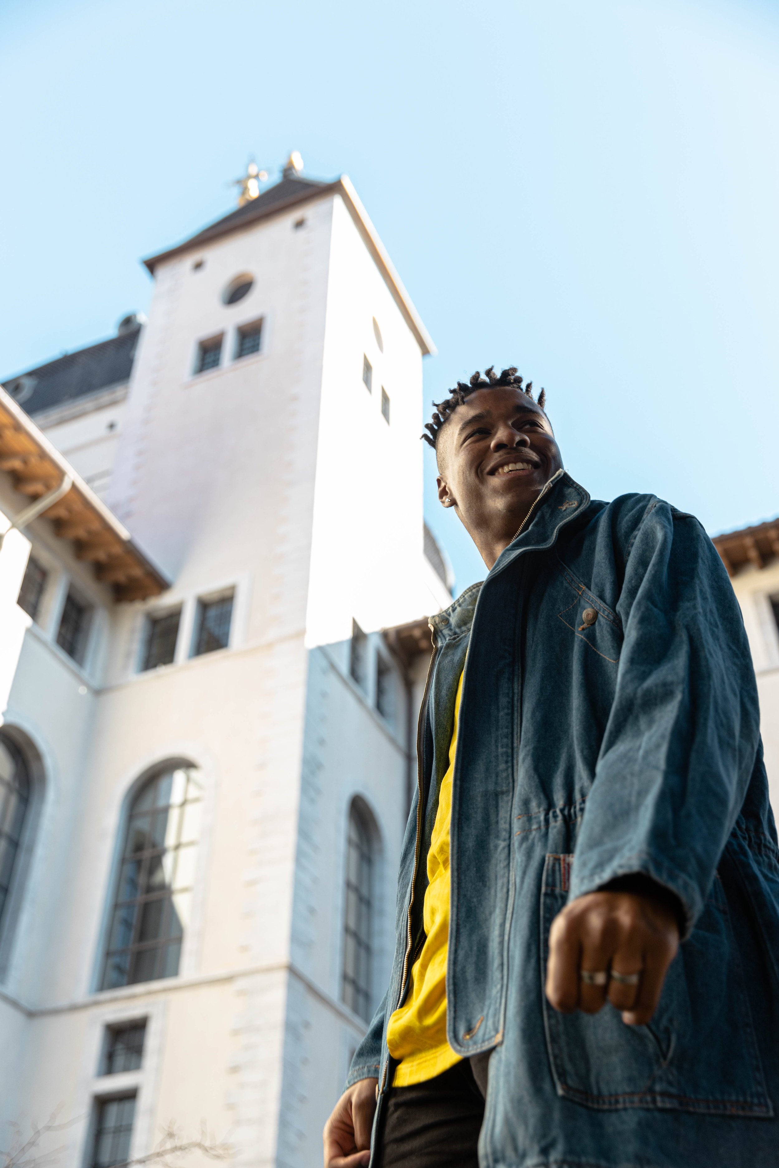 Jeune homme souriant portant une veste en jean, debout devant un bâtiment blanc avec une tour, ciel bleu en arrière-plan.