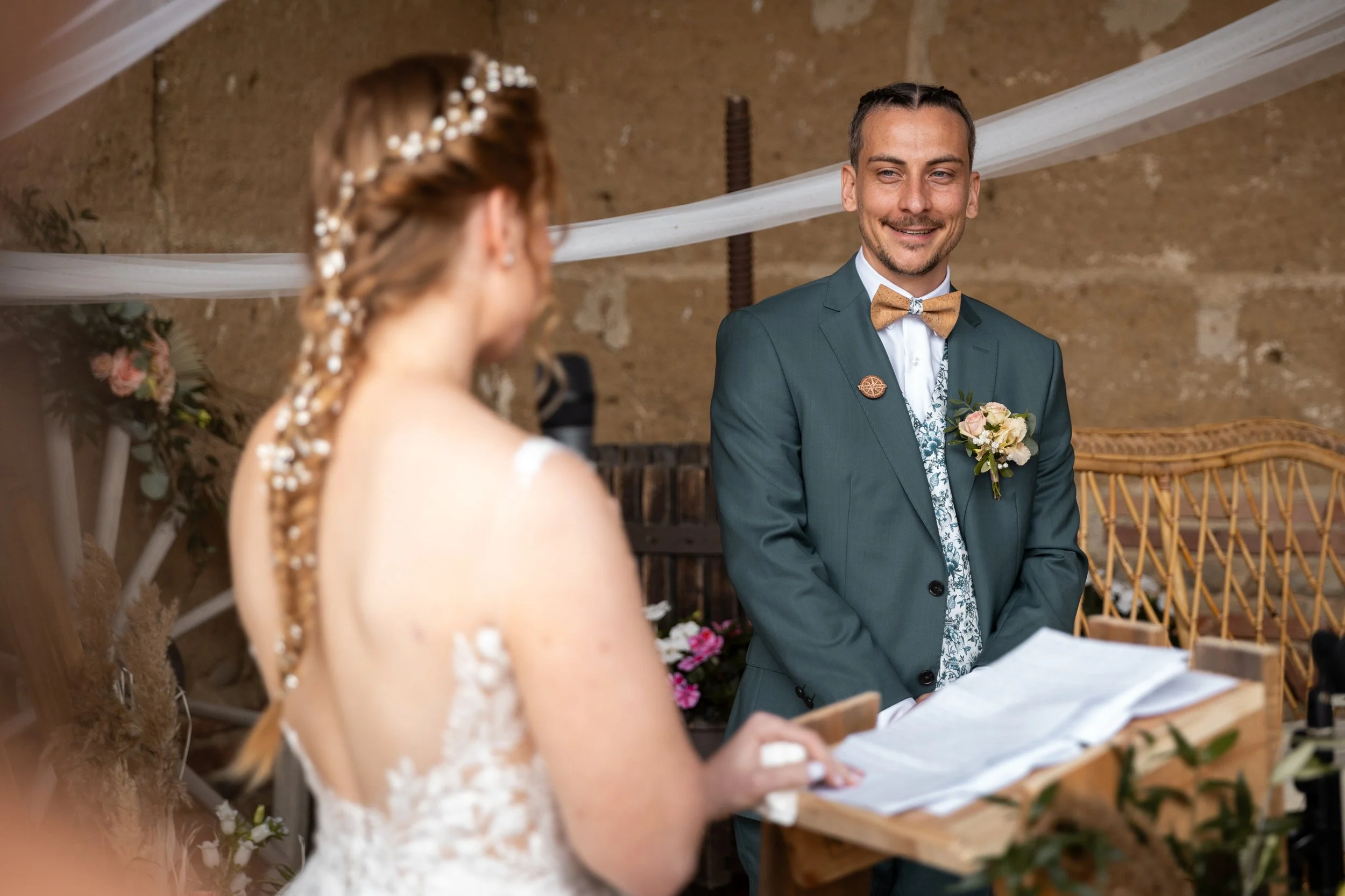 Un homme souriant en costume lors d'un mariage, regardant une femme dont le visage n'est pas visible, dans une décoration rustique avec des fleurs et des tissus blancs.