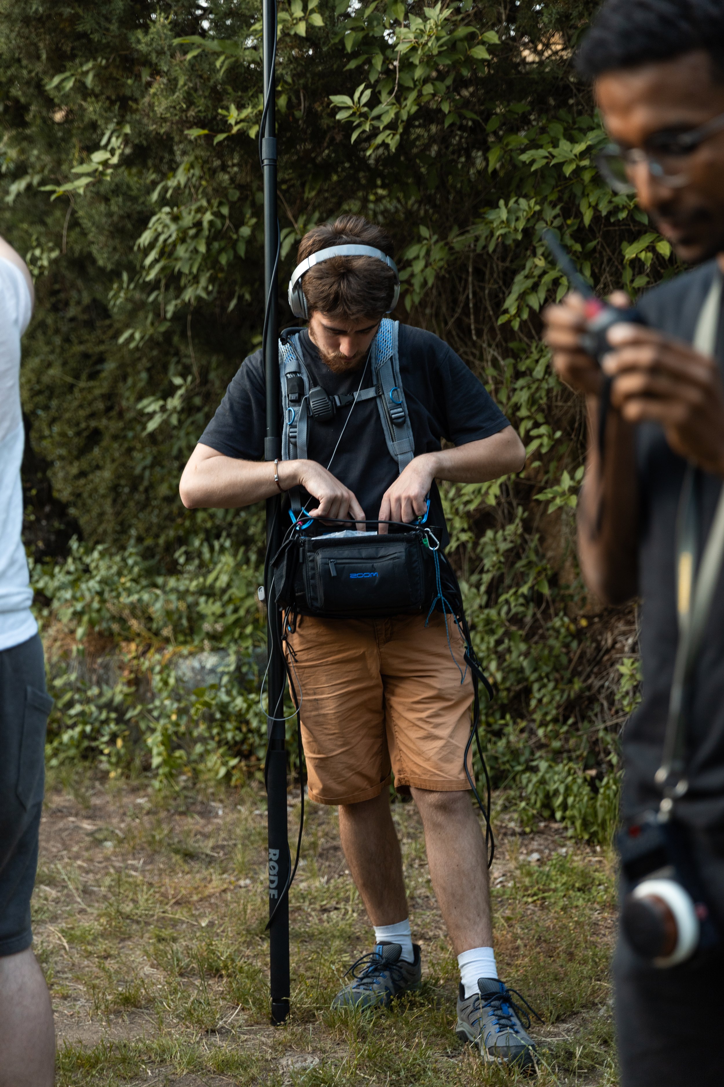 Un jeune homme avec un casque audio, une sacoche et une caméra en outdoor, probablement en train de préparer de l'équipement ou de régler quelque chose.