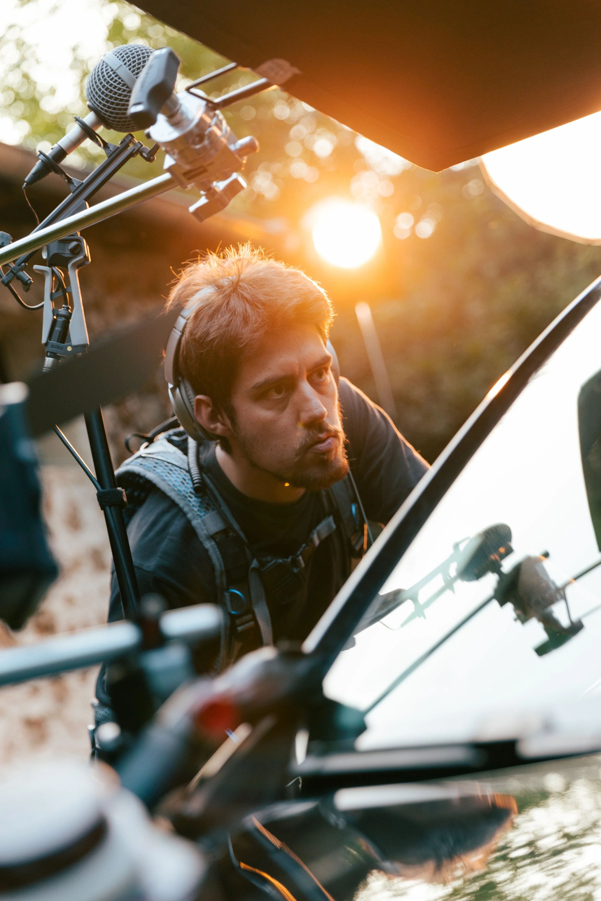 Un homme avec un casque d'écoute regarde un écran lors d'une séance de tournage en plein air avec le soleil couchant en arrière-plan.