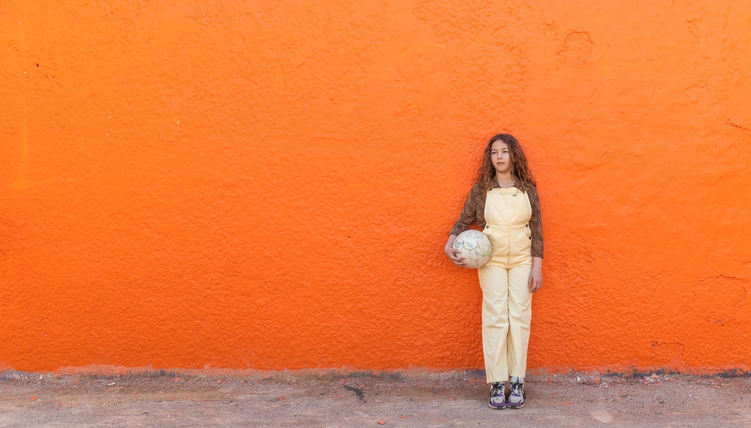 Jeune fille avec des cheveux longs et bouclés, portant une combinaison beige, tenant un ballon de foot, debout devant un mur orange.