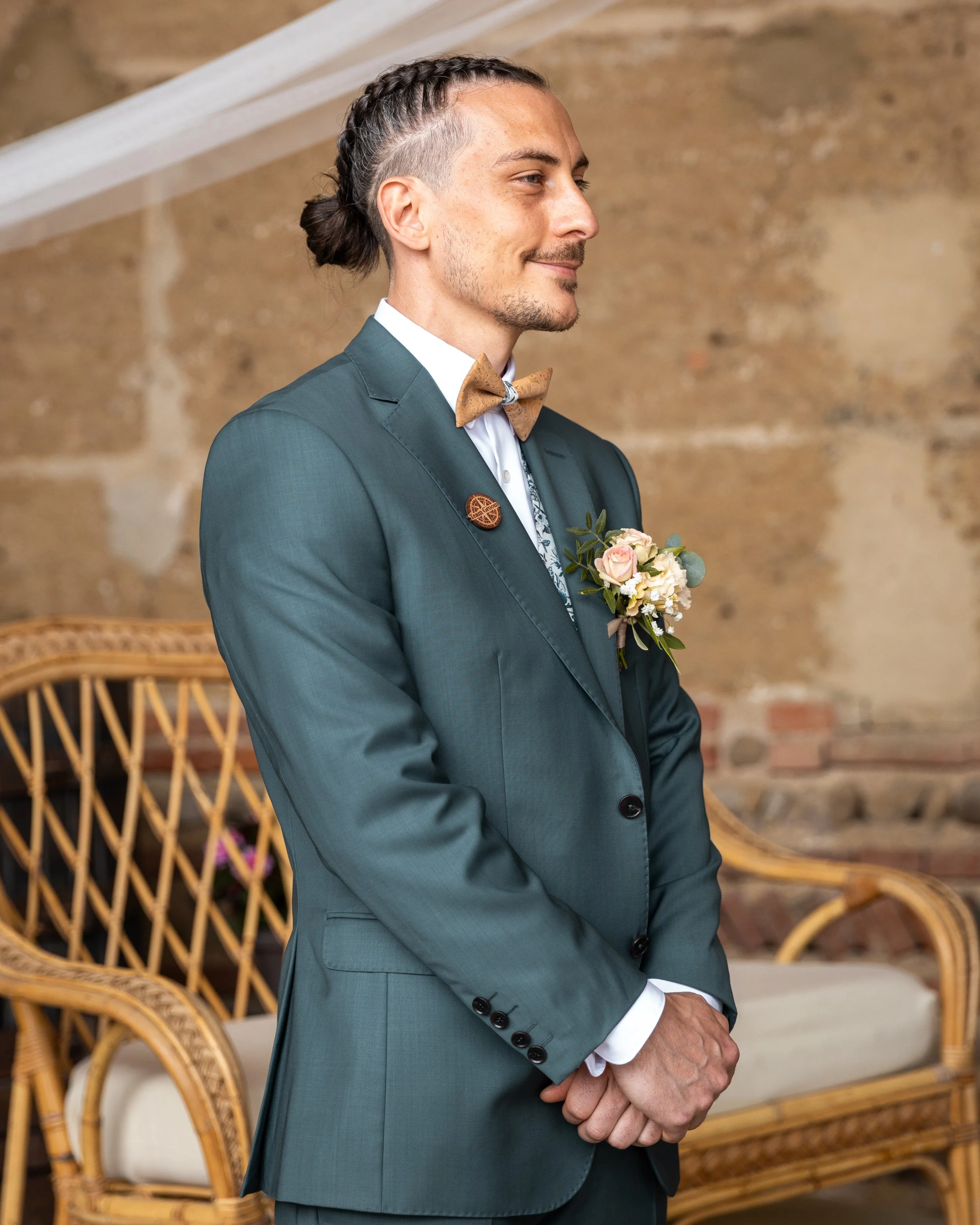 Jeune homme en costume vert, avec un nœud papillon beige, portant une boutonnière florale, lors d'une cérémonie de mariage, debout avec les mains croisées, dans une pièce avec un canapé en rotin et un mur en briques.
