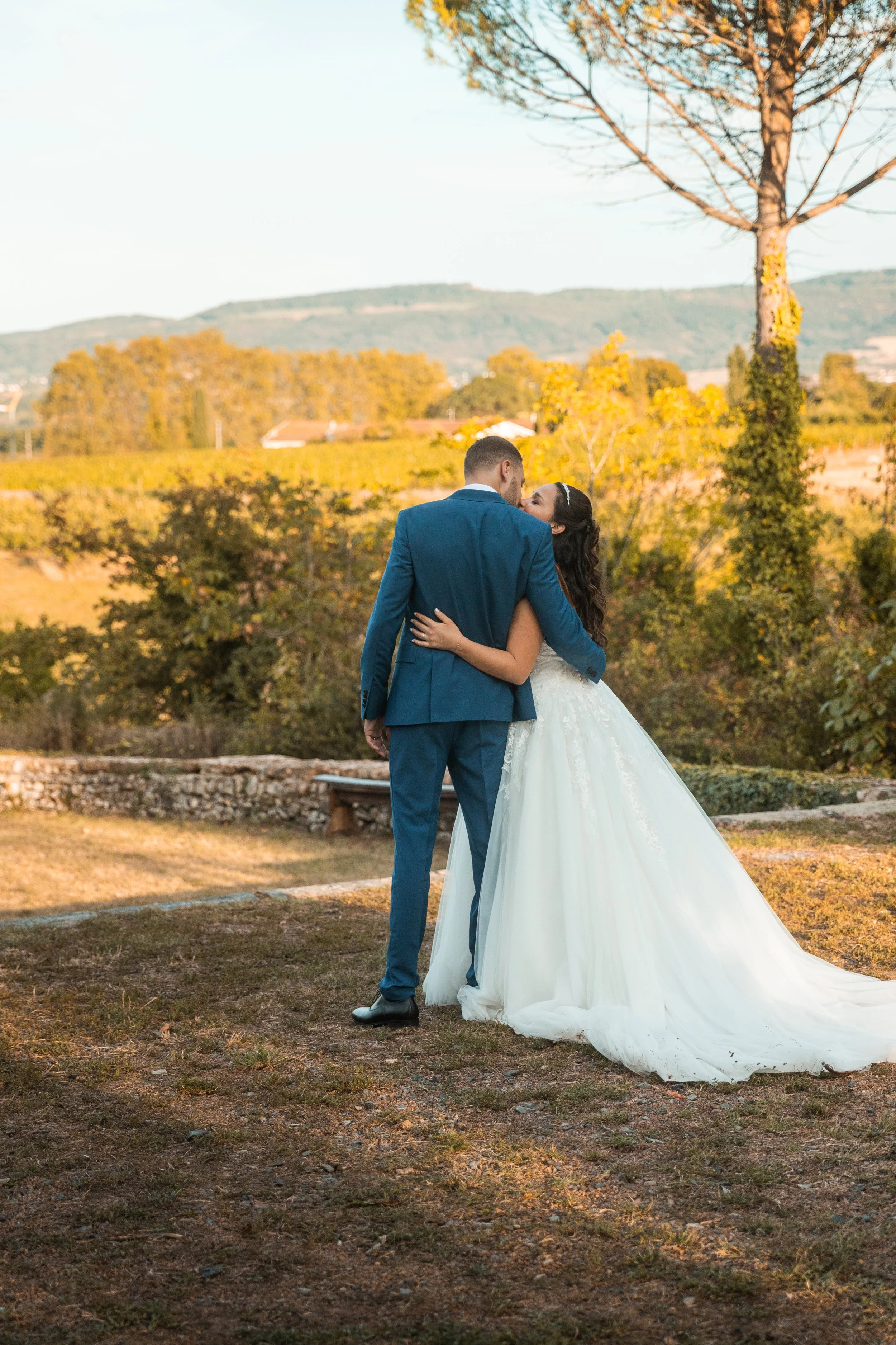 Un couple de mariés se tenant par la taille, partagé un baiser dans un décor naturel et champêtre, avec des arbres et des collines en arrière-plan, lors d'une journée ensoleillée.