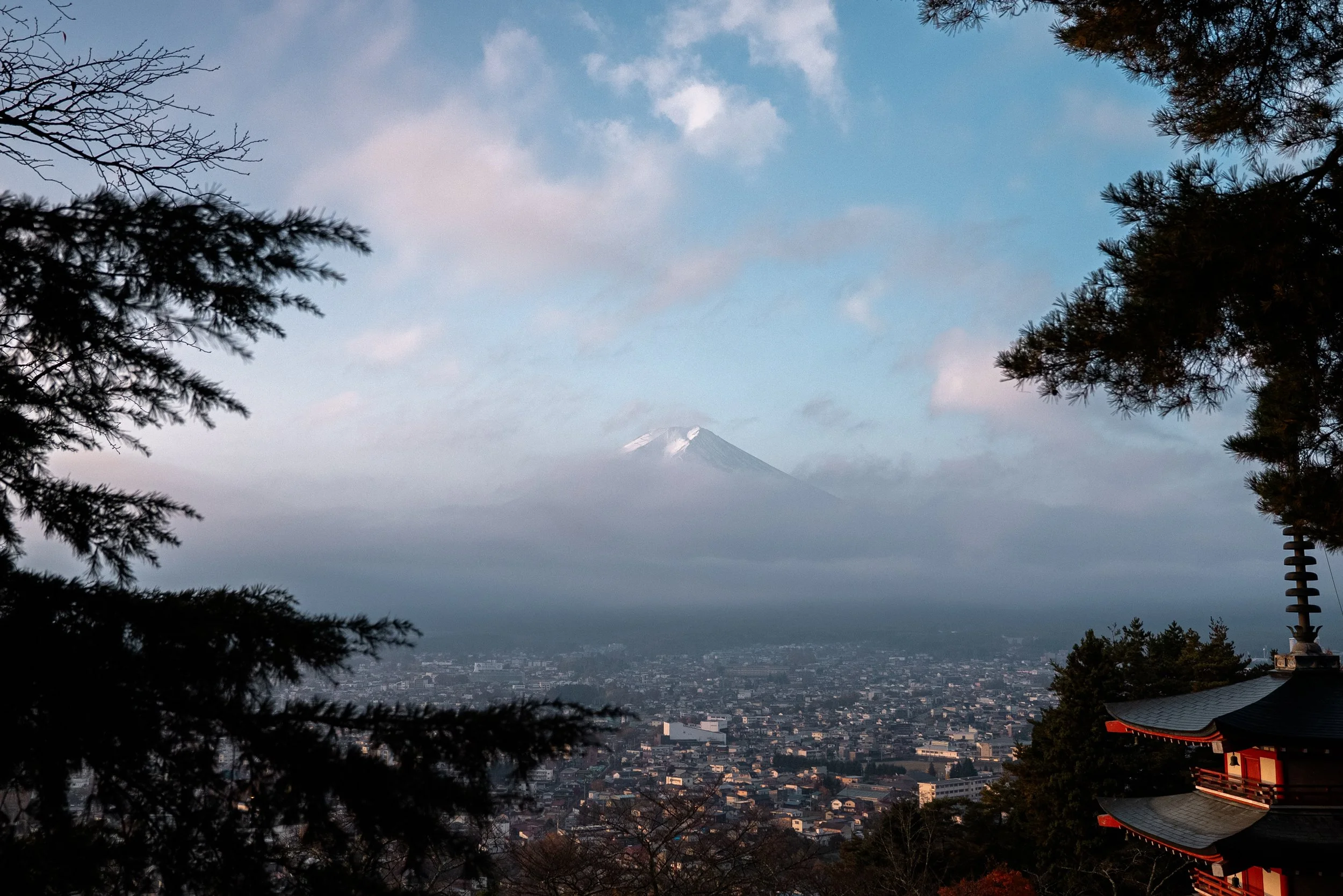 Une vue panoramique de Tokyo avec la ville en premier plan, entourée d'arbres, et le mont Fuji enneigé en arrière-plan, partiellement couvert de nuages.