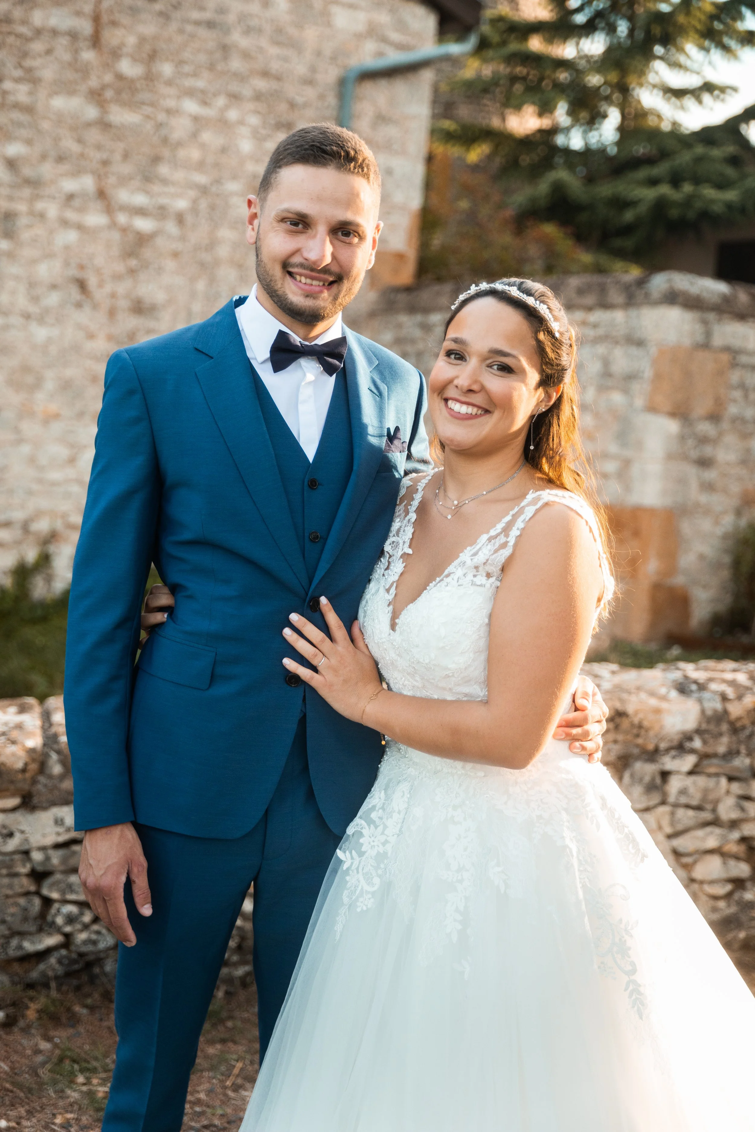 Un couple de mariés posa pour une photo en extérieur, la femme porte une robe de mariage blanche et le homme un costume bleu avec un nœud papillon, dans un cadre rustique avec un mur en pierre en arrière-plan.