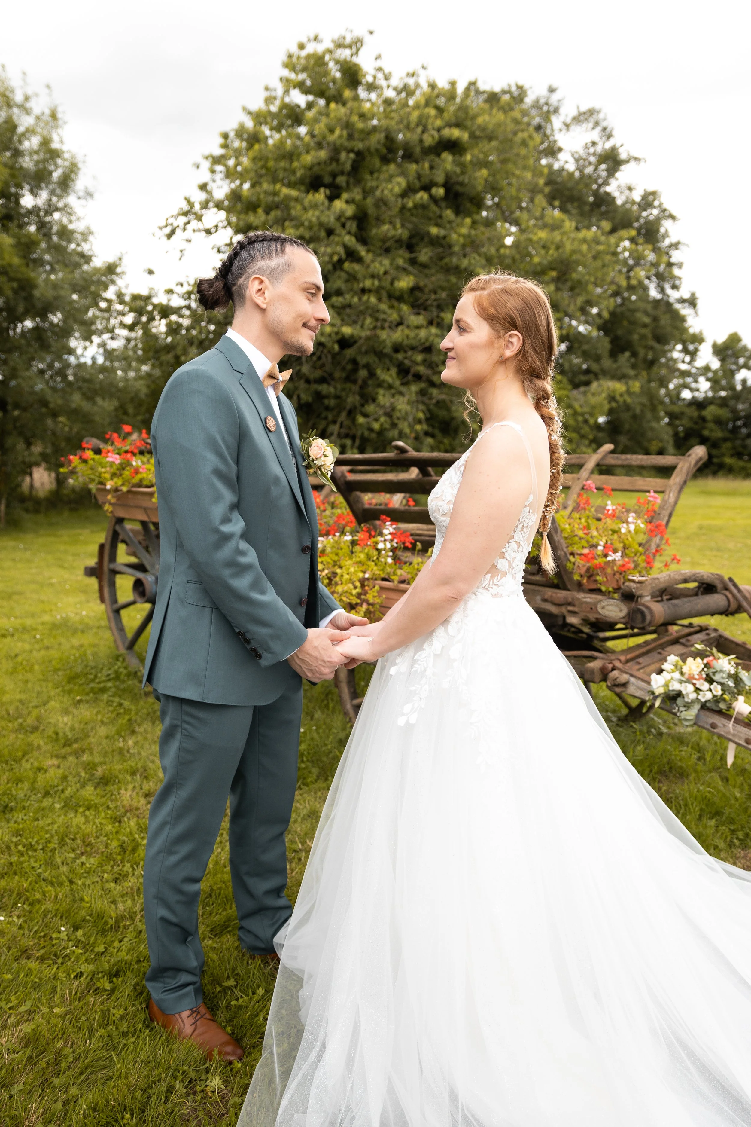 Un couple de mariés qui se tient la main lors de leur mariage en extérieur, avec un arrière-plan de chariot en bois et de fleurs.