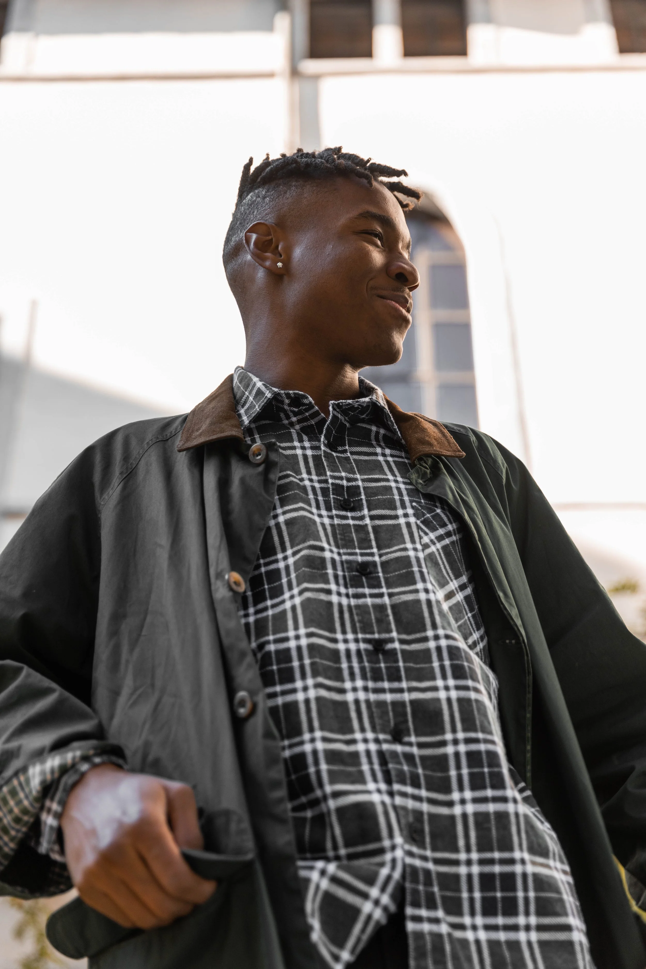 Jeune homme avec dreadlocks, portant une chemise à carreaux et une veste en cuir, souriant.