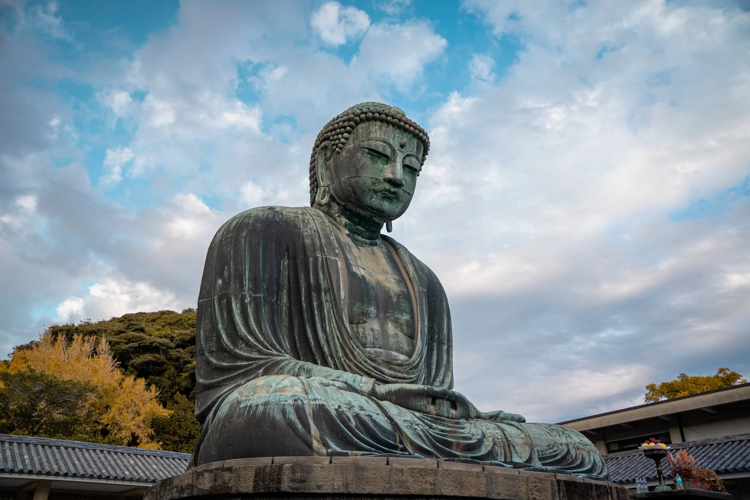 Gros Bouddha en bronze assis dans un temple, avec un ciel nuageux en arrière-plan.