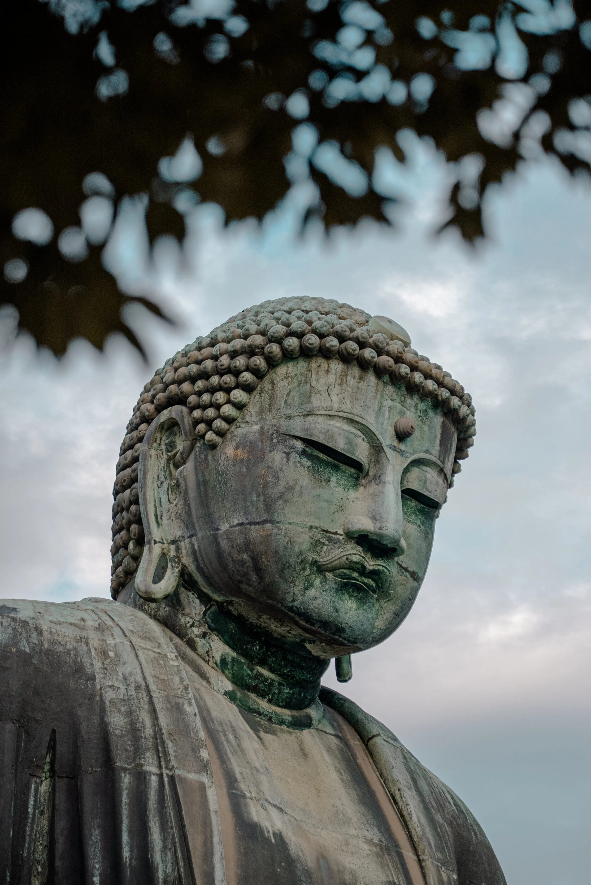 Une statue en bronze d'un Bouddha avec un visage serein, des yeux mi-clos et des cheveux en boucles, avec un ciel nuageux en arrière-plan