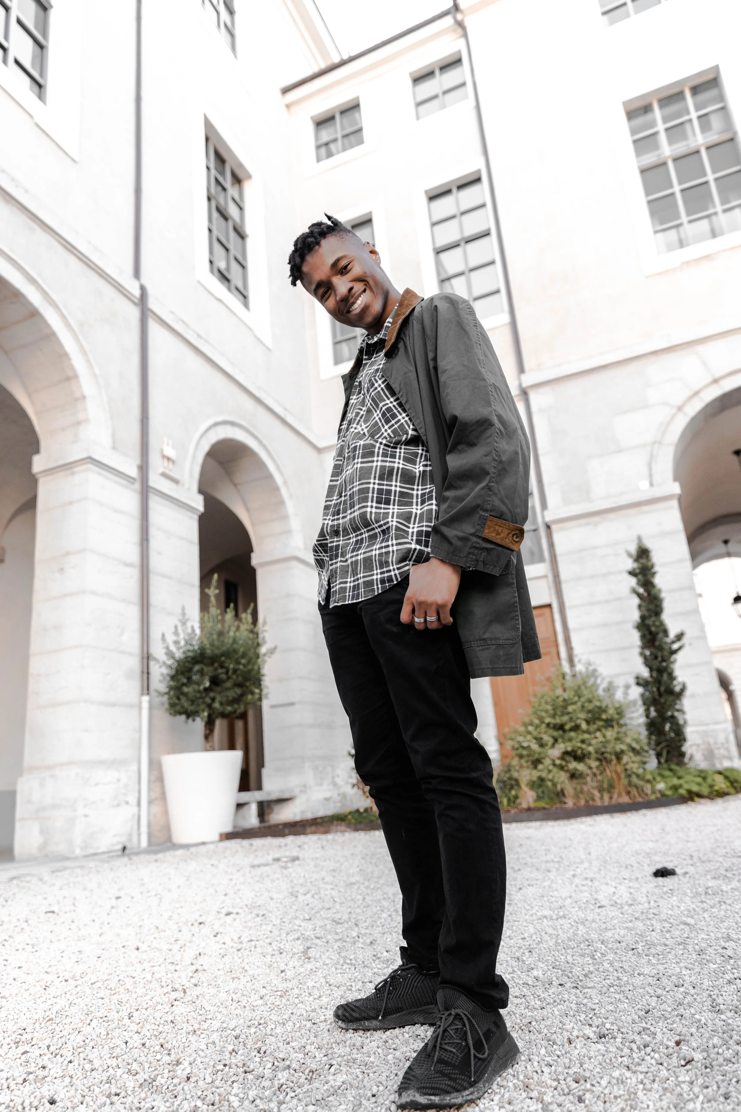 Jeune homme souriant debout dans une cour pavée devant un bâtiment blanc avec des arches et des fenêtres, portant une chemise à carreaux, une veste noire, et des chaussures de sport noires.