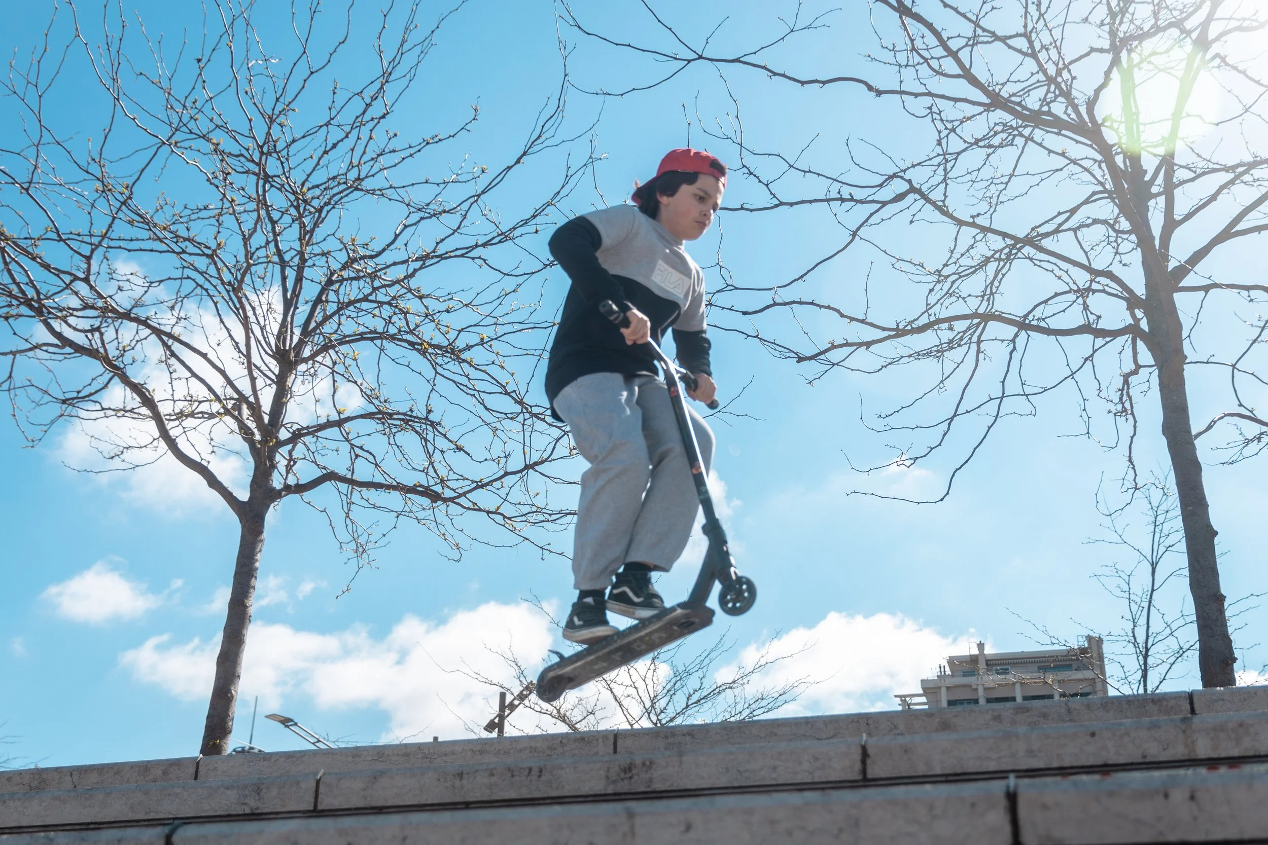 Jeune garçon faisant du scooter électrique dans un parc par une journée ensoleillée, sans feuilles sur les arbres.