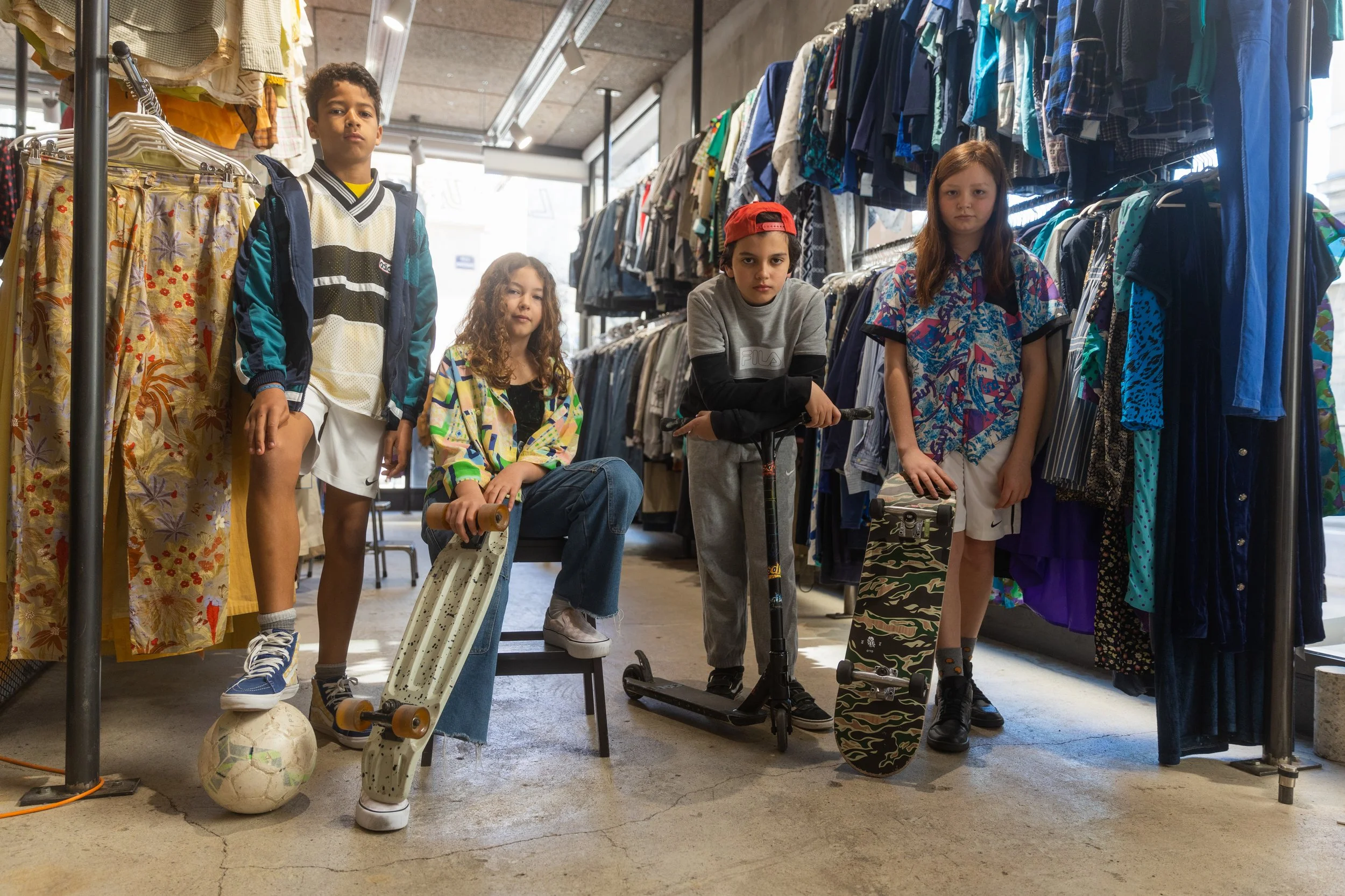 Cinq enfants dans une boutique de vêtements et de skateboards, portant des vêtements décontractés et tenant ou posant avec leurs skateboards, parmi des rayons de vêtements.