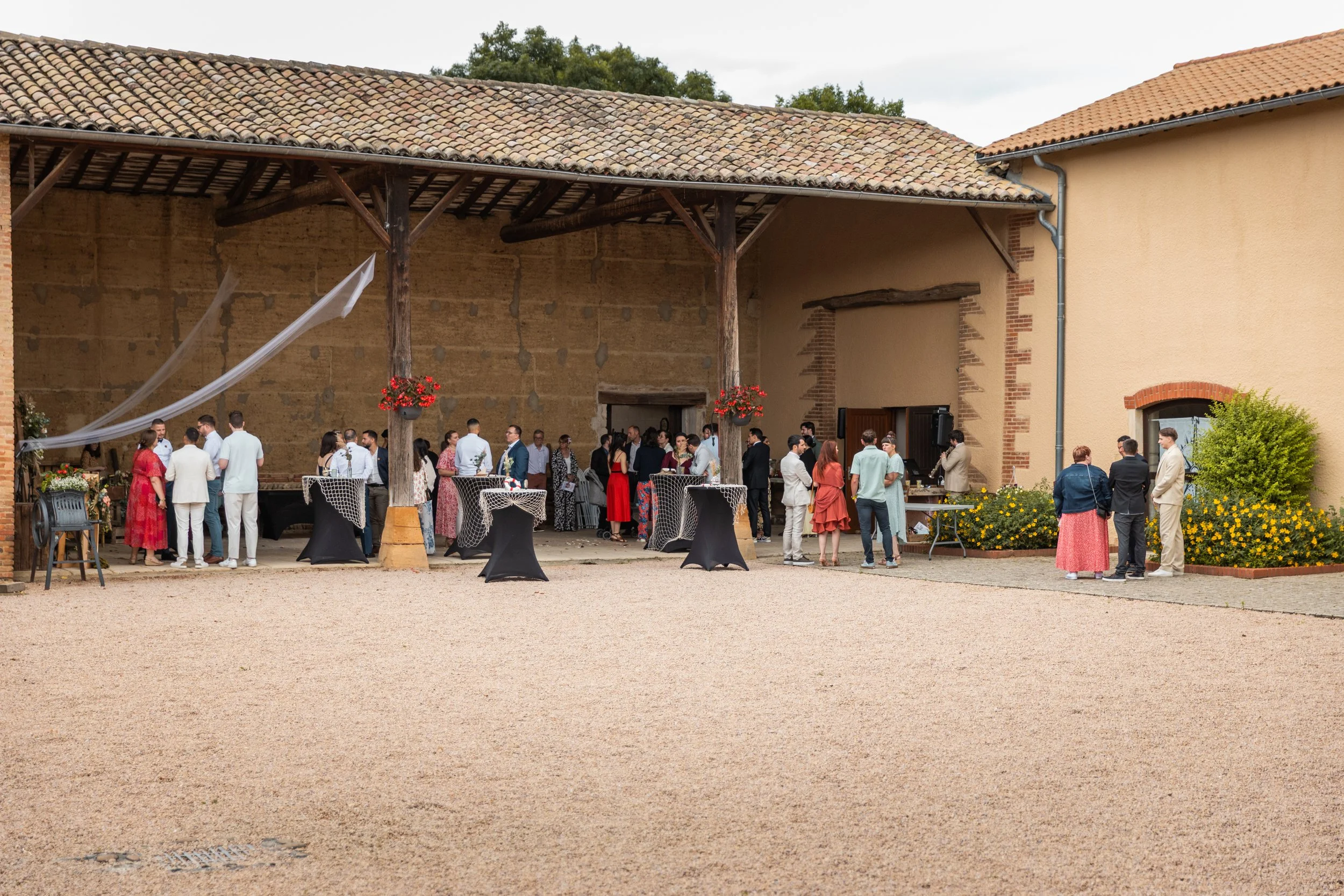 Un groupe de personnes lors d'une fête en plein air, sous un grand bâtiment en pierre avec un toit en tuiles, décoré de fleurs rouges et d'une toile blanche, avec des tables hautes et un barbecue.