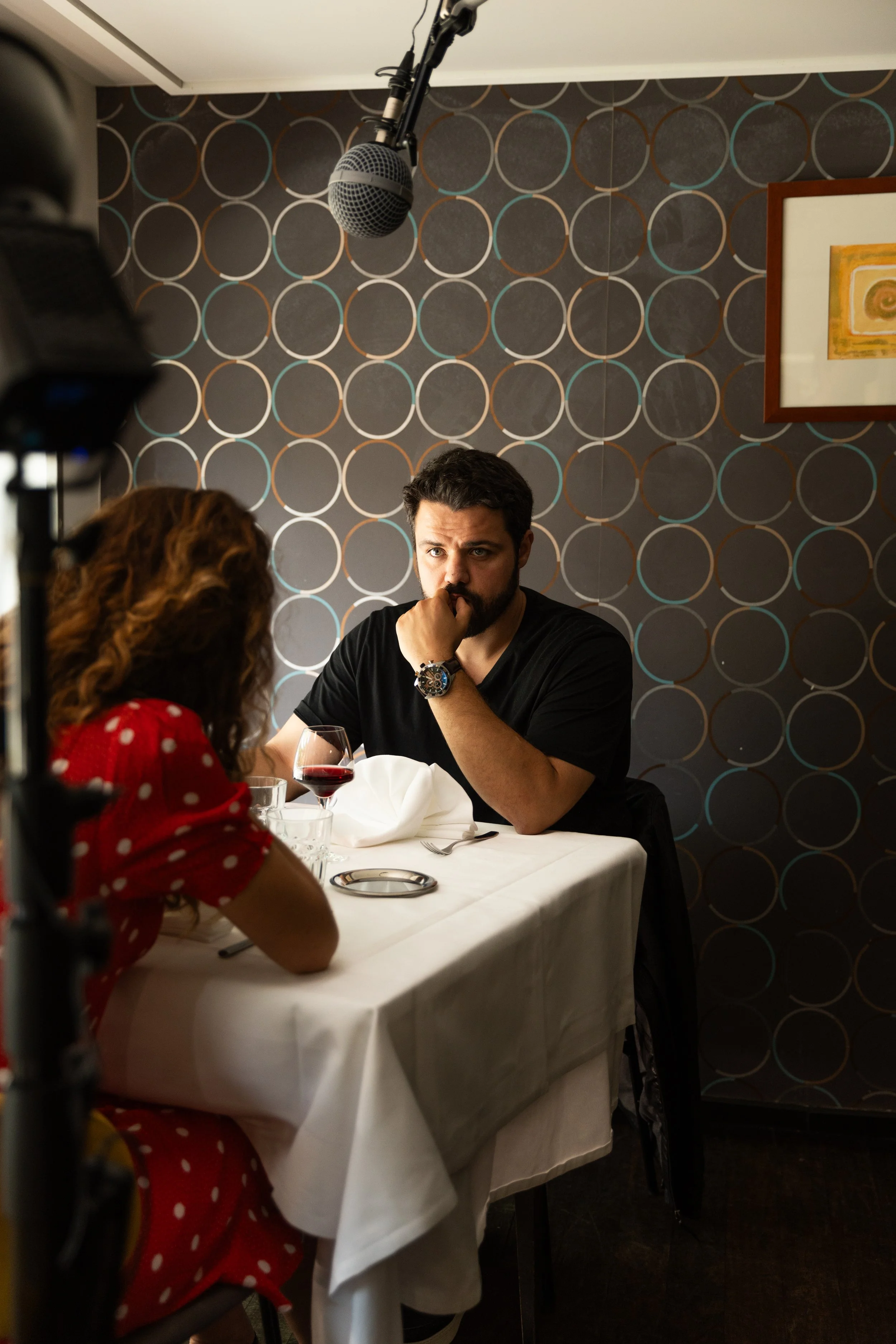 Un homme et une femme assis à une table dans un restaurant, parlant. La femme porte une robe rouge à pois blancs, et il y a un verre de vin rouge sur la table. La scène semble être une prise de vue pour un projet vidéo ou photo.