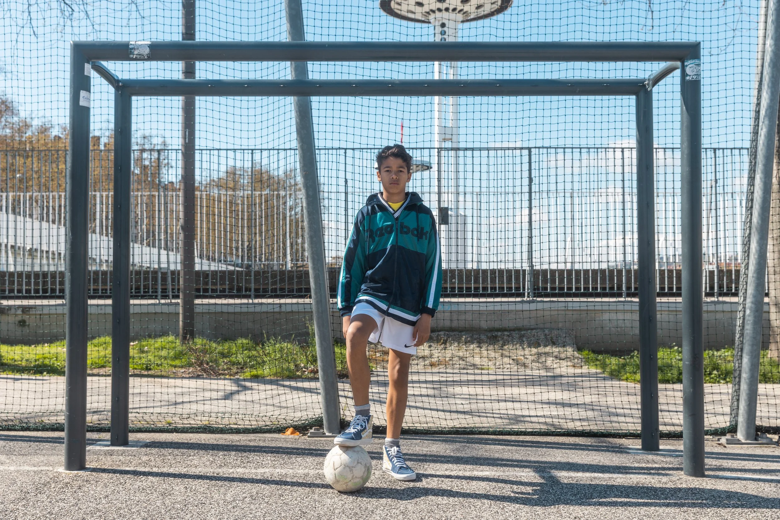 Jeune garçon en tenue de sport, debout devant un but de football, avec un ballon à ses pieds, en plein air, ciel bleu.