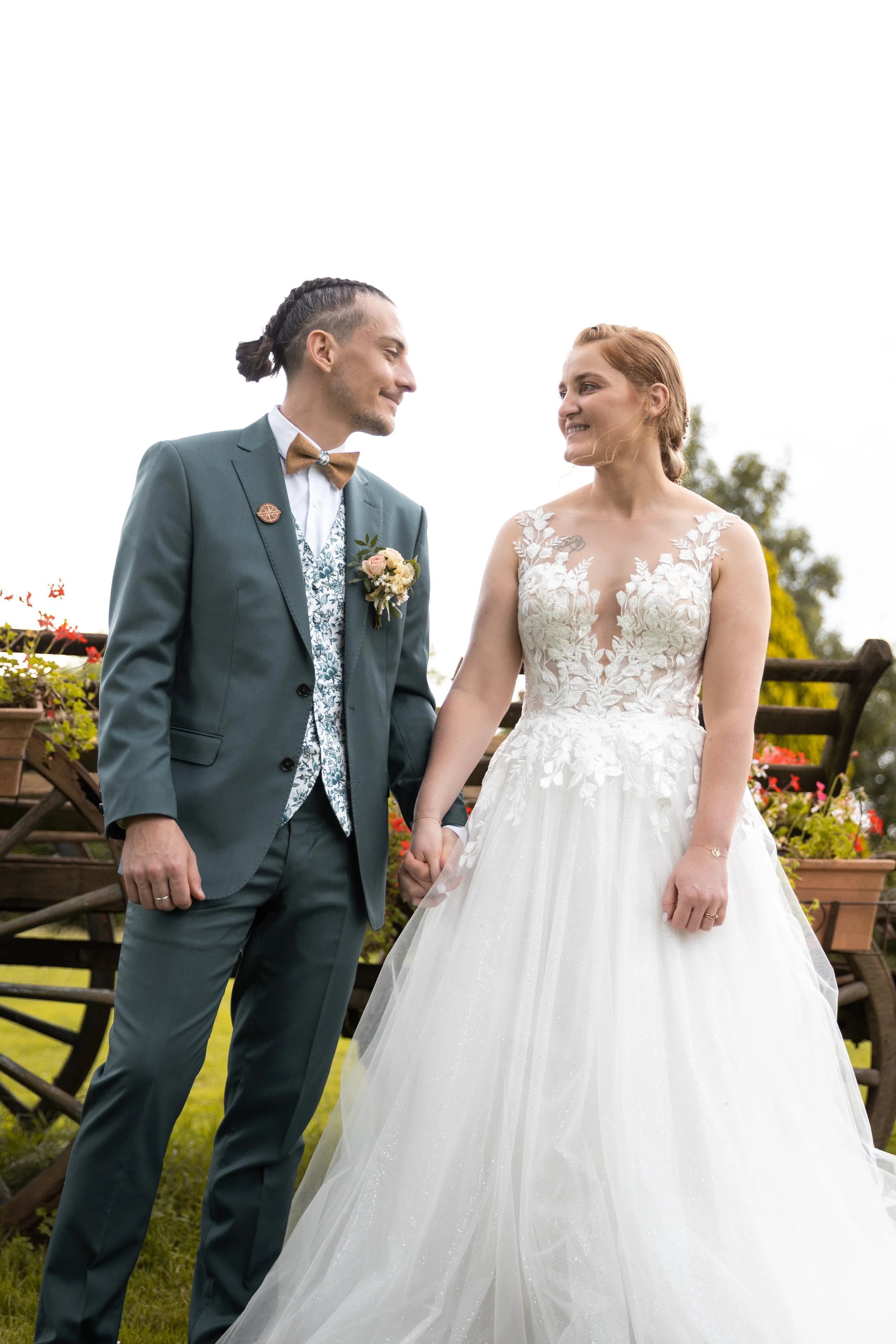 Un couple de mariés se tient dans un jardin, elle en robe de mariage blanche et lui en costume gris avec un nœud papillon