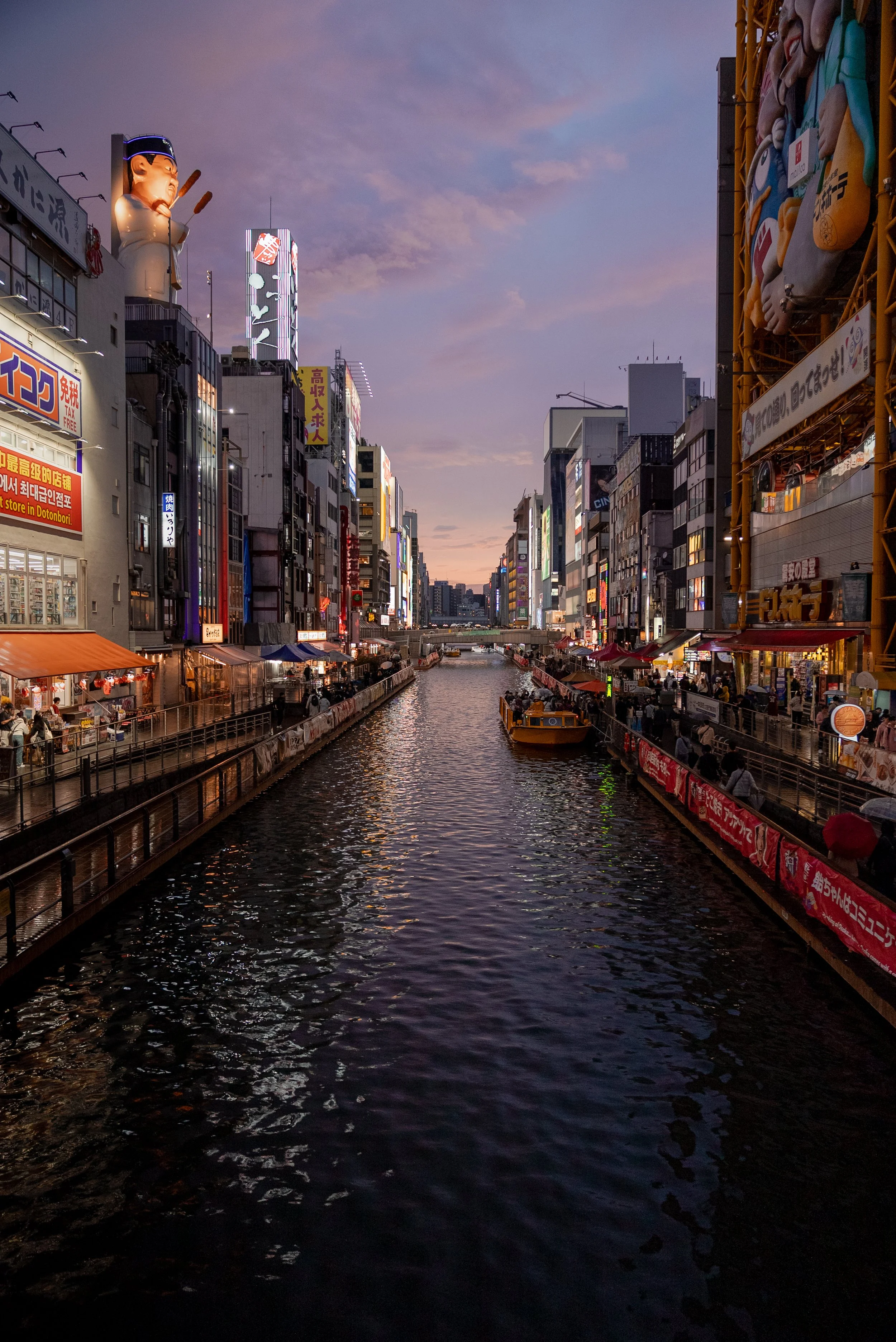 Vue d'une rivière urbaine bordée de bâtiments lumineux la nuit, avec des passants et des bateaux sur l'eau, dans une ville animée.