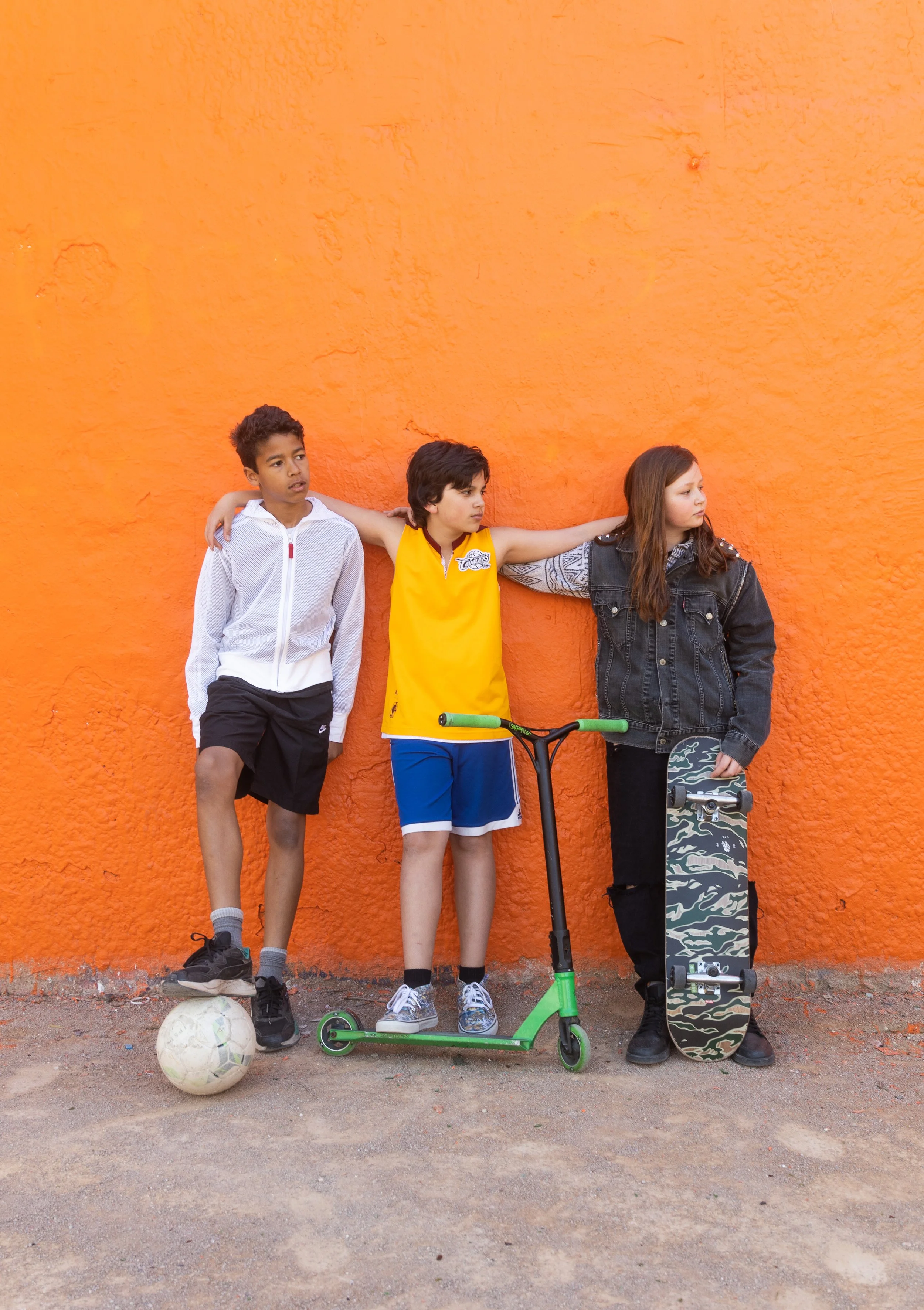 Trois enfants posent contre un mur orange, avec un ballon de football, une trottinette et une planche à roulettes.