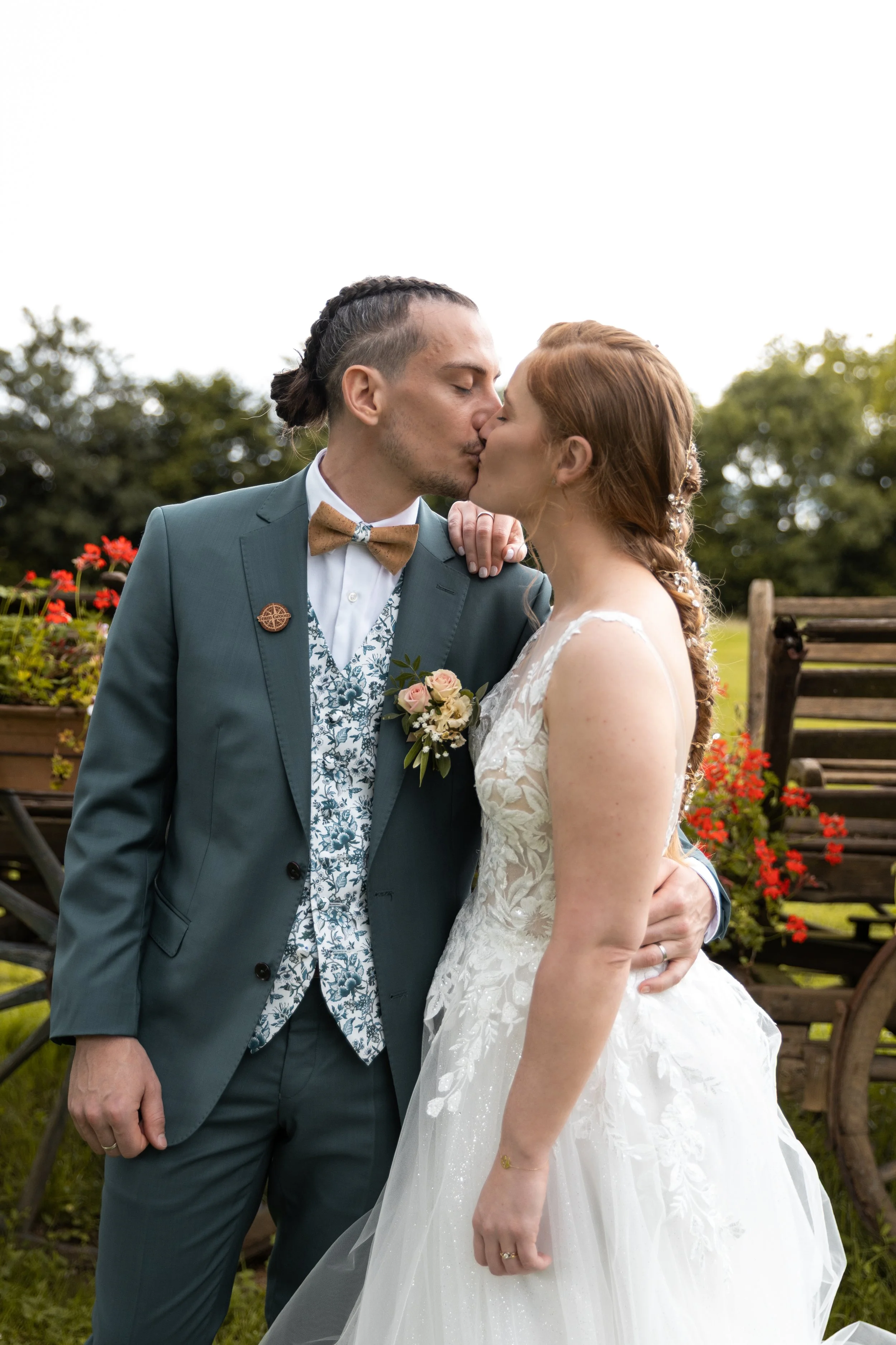 Un couple en robe de mariage s'embrasse, dans un jardin avec des fleurs et des arbres.