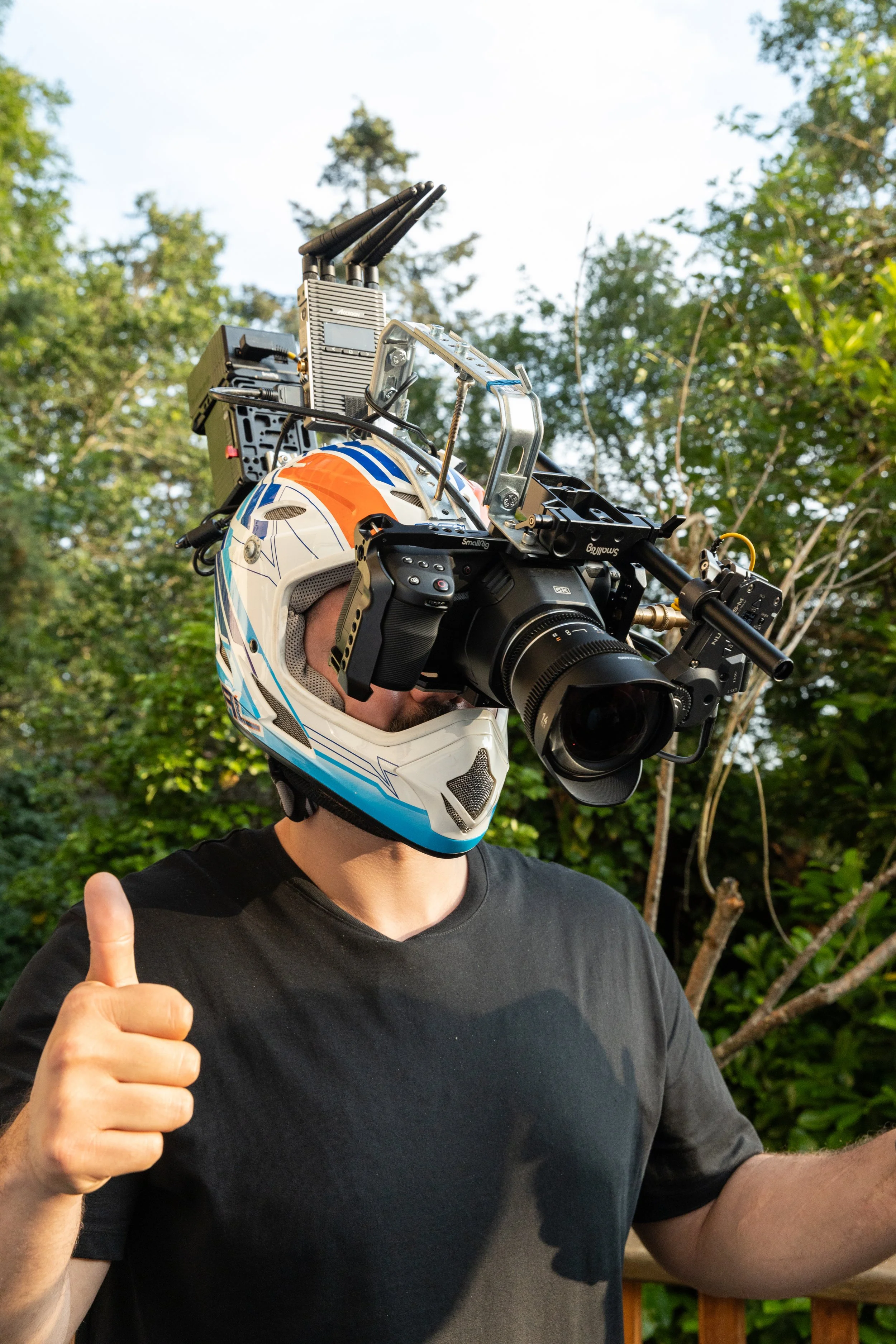 Une personne porte un casque de moto et un système de caméra complexe, faisant un signe de pouce en l'air dans un environnement extérieur verdoyant.