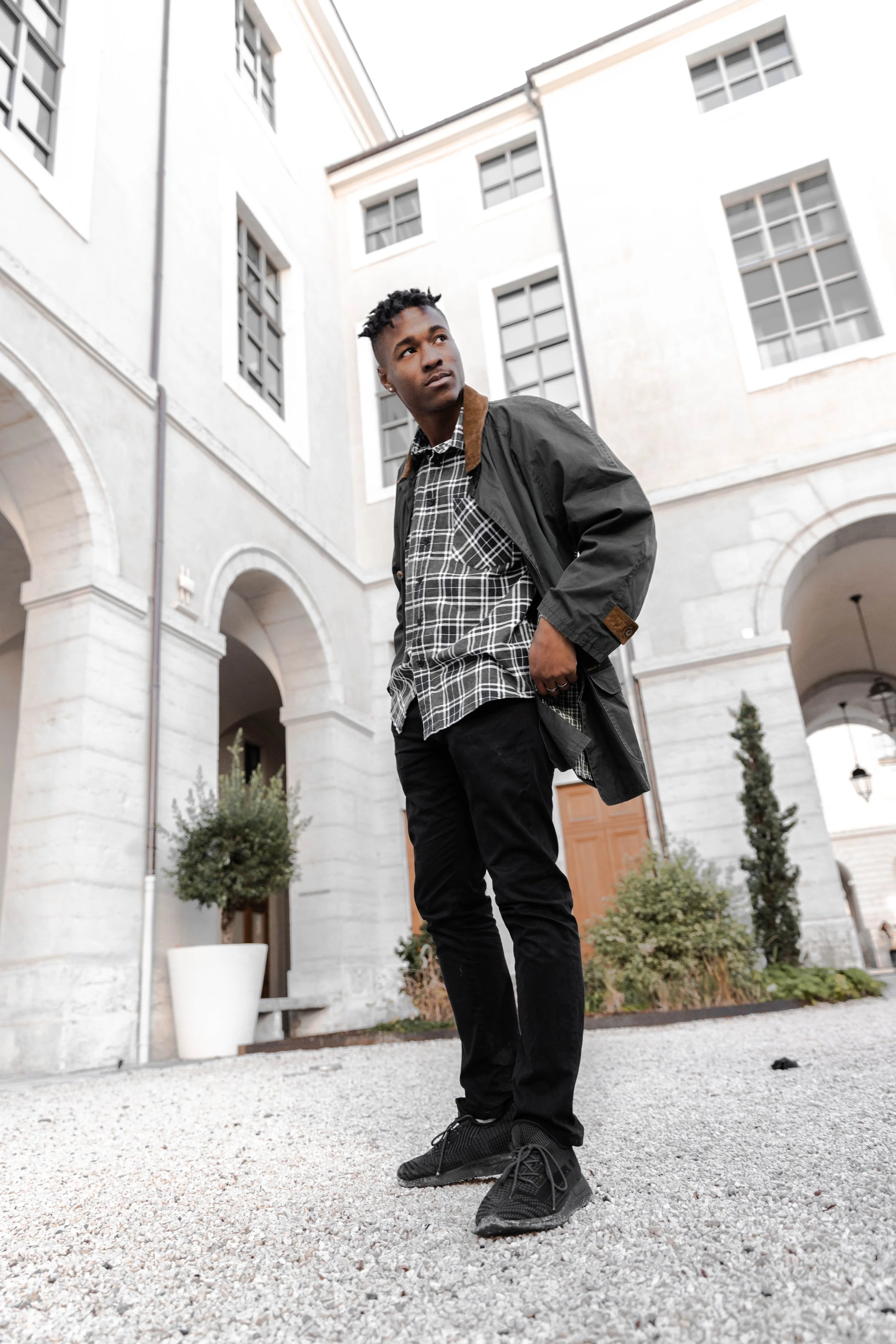Un jeune homme debout dans une cour pavée, portant un manteau noir, une chemise à carreaux, et des chaussures noires, avec un bâtiment blanc en arrière-plan.