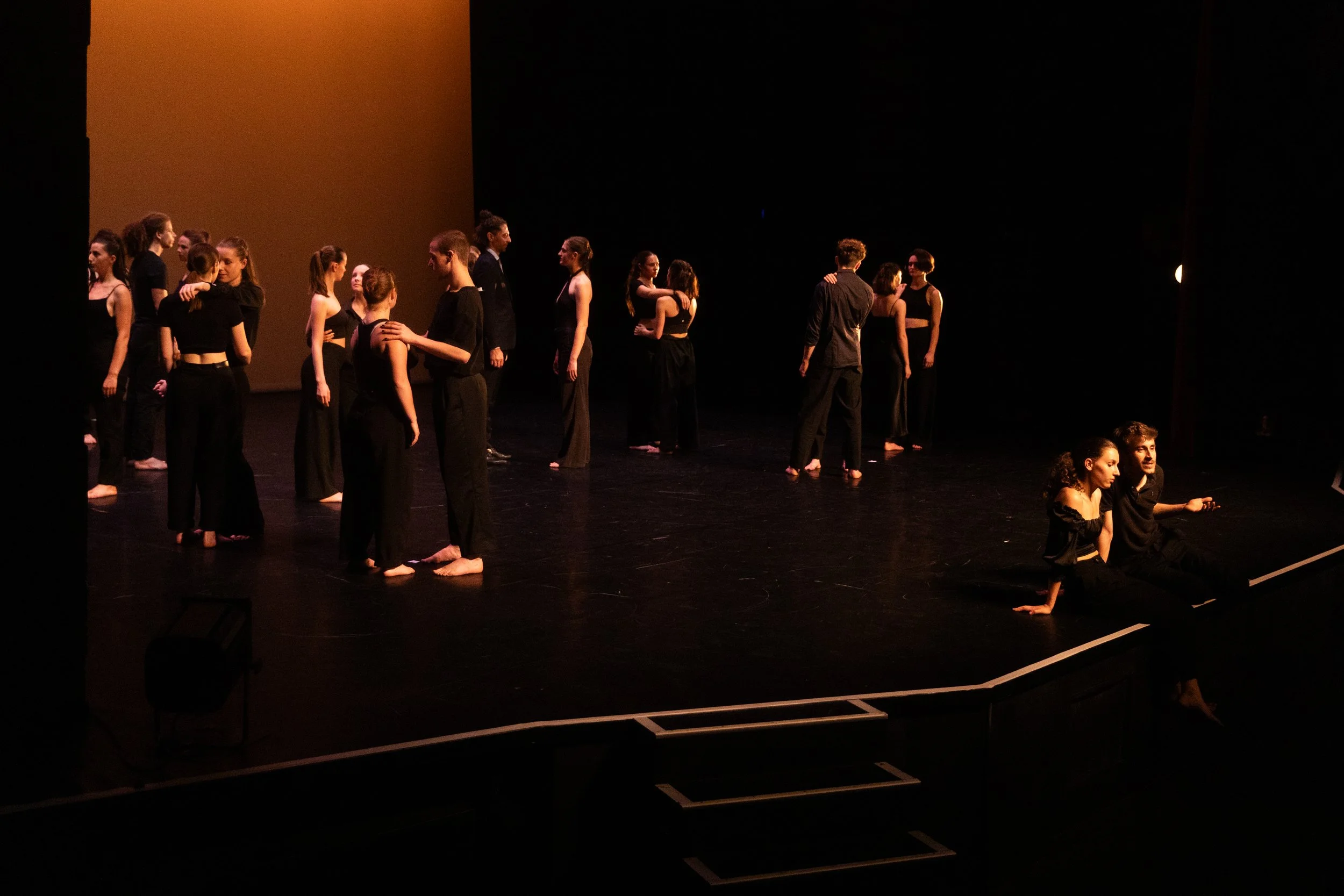 Groupe de jeunes artistes sur scène lors d'une représentation de danse ou de théâtre, certains en duo, d'autres en groupe, avec un décor simple et lumière tamisée.
