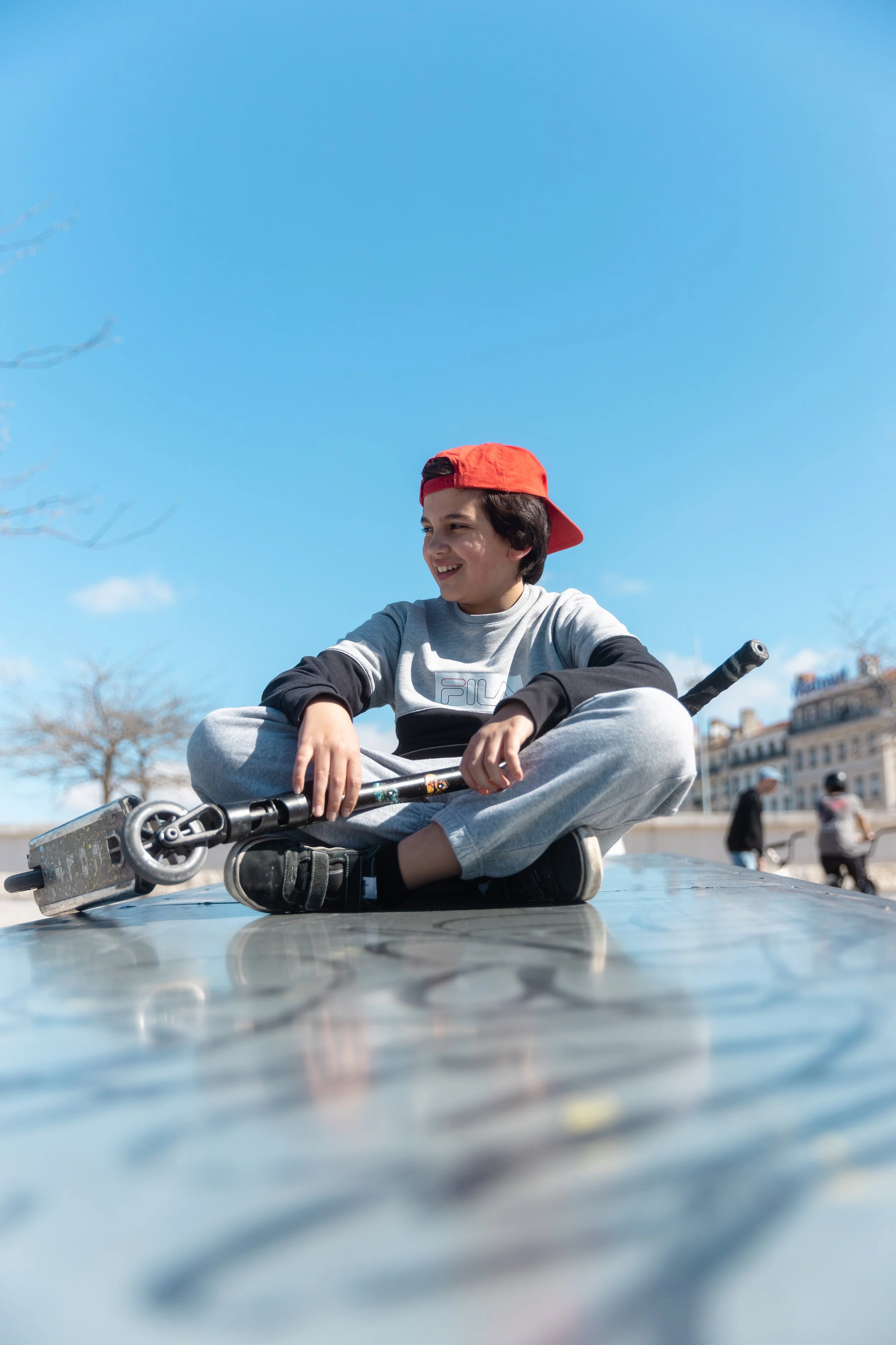 Un jeune garçon assis croisé, portant un sweat-shirt gris, un jean gris, un chapeau rouge et des chaussures noires, souriant, tenant un trottinette électrique sur une surface métallique en plein air, avec un ciel bleu clair et des bâtiments en arrièr