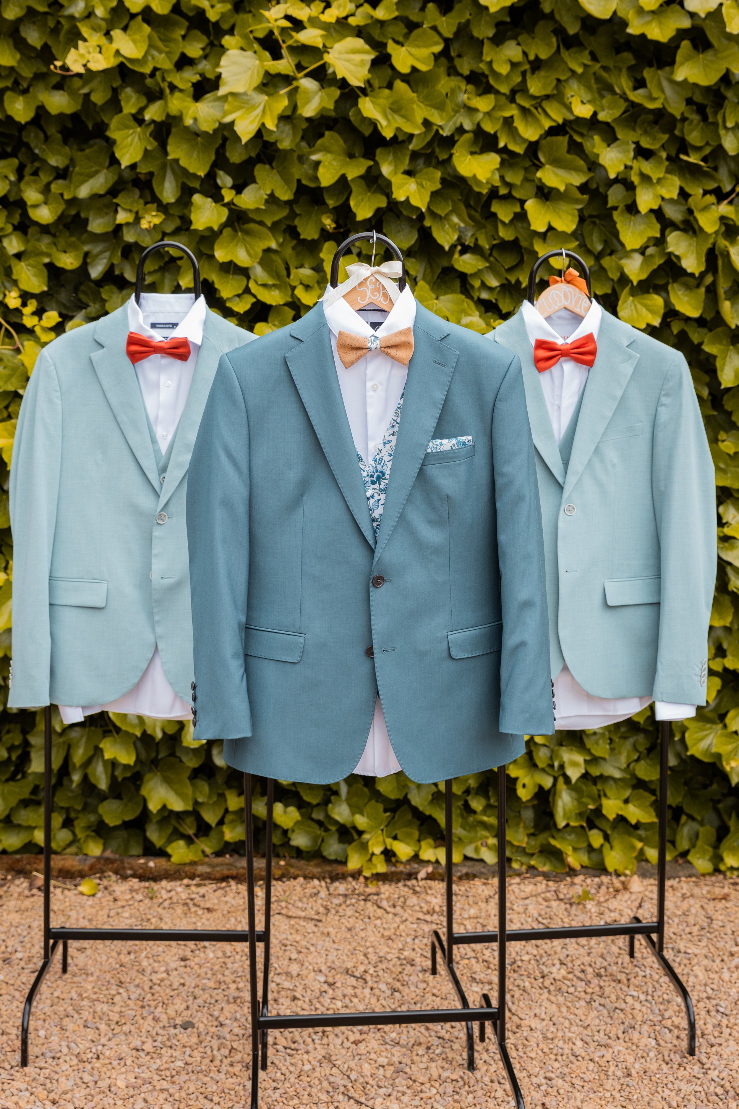 Costumes de mariage en costume bleu clair avec nœuds papillon rouges ou beige, suspendus sur des portants en extérieur avec un mur de feuilles vertes en arrière-plan.