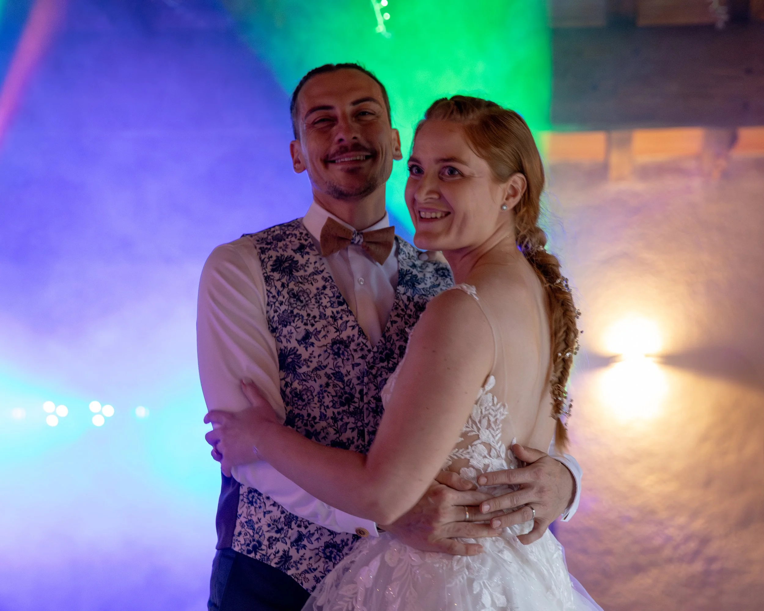 Un couple danse lors de leur mariage, la femme porte une robe blanche et l'homme un veston avec un nœud papillon beige, sous un éclairage coloré.