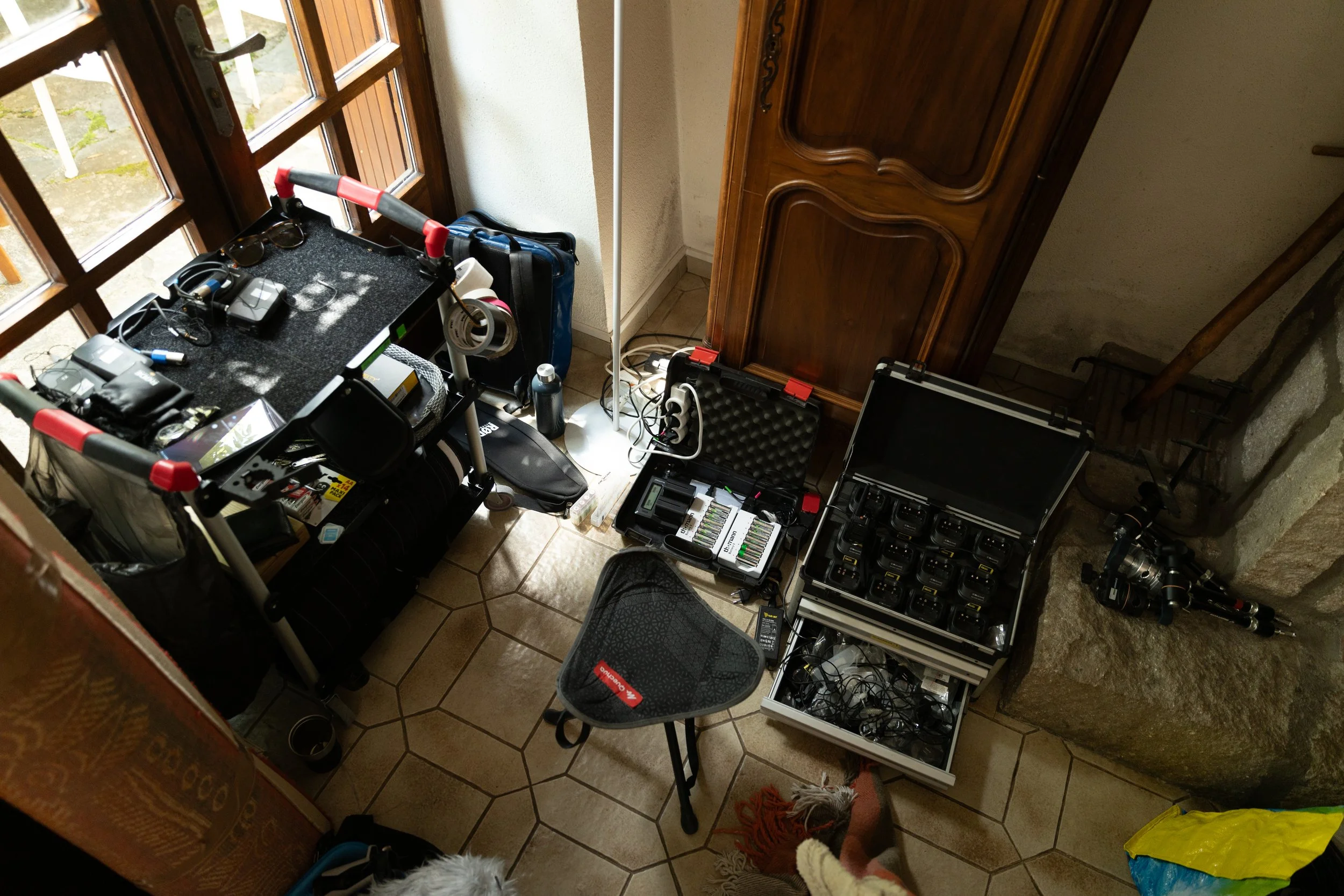 Une pièce avec du matériel de photographie et de vidéos, comprenant des caméras, des batteries, des câbles, une table avec divers appareils et accessoires, un trépied, une chaise pliable, et un coffre de rangement, près d'une porte-fenêtre en bois.