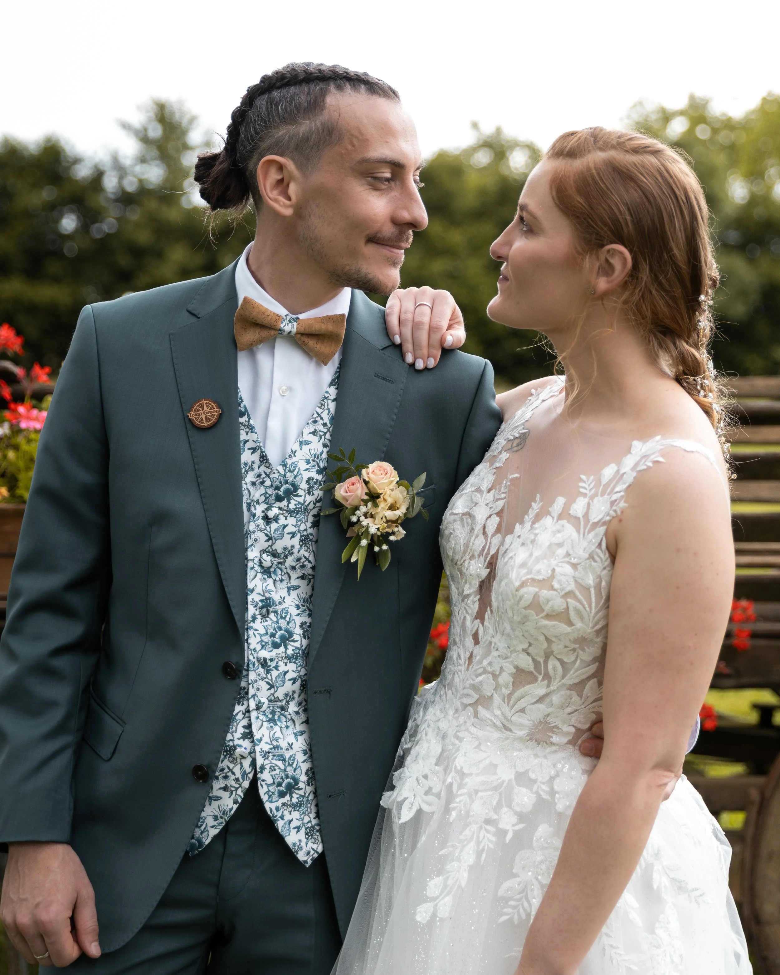 Un couple lors d'un mariage, l'homme porte un costume bleu avec un nœud papillon en velours et un bouton de marié, la femme porte une robe de mariée blanche avec broderies florales.