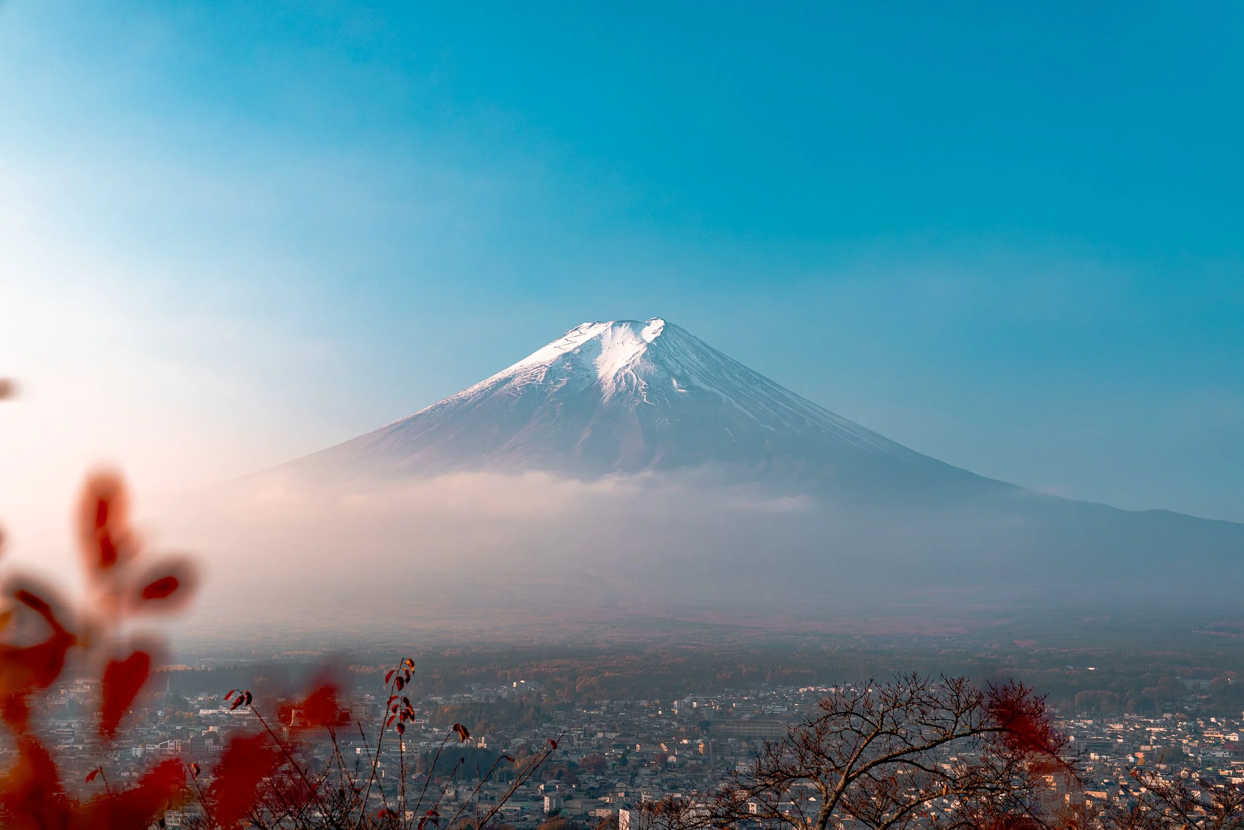 Photographie du Mont Fuji avec des nuages autour de sa base, vue d'une ville au premier plan, ciel clair et ensoleillé.