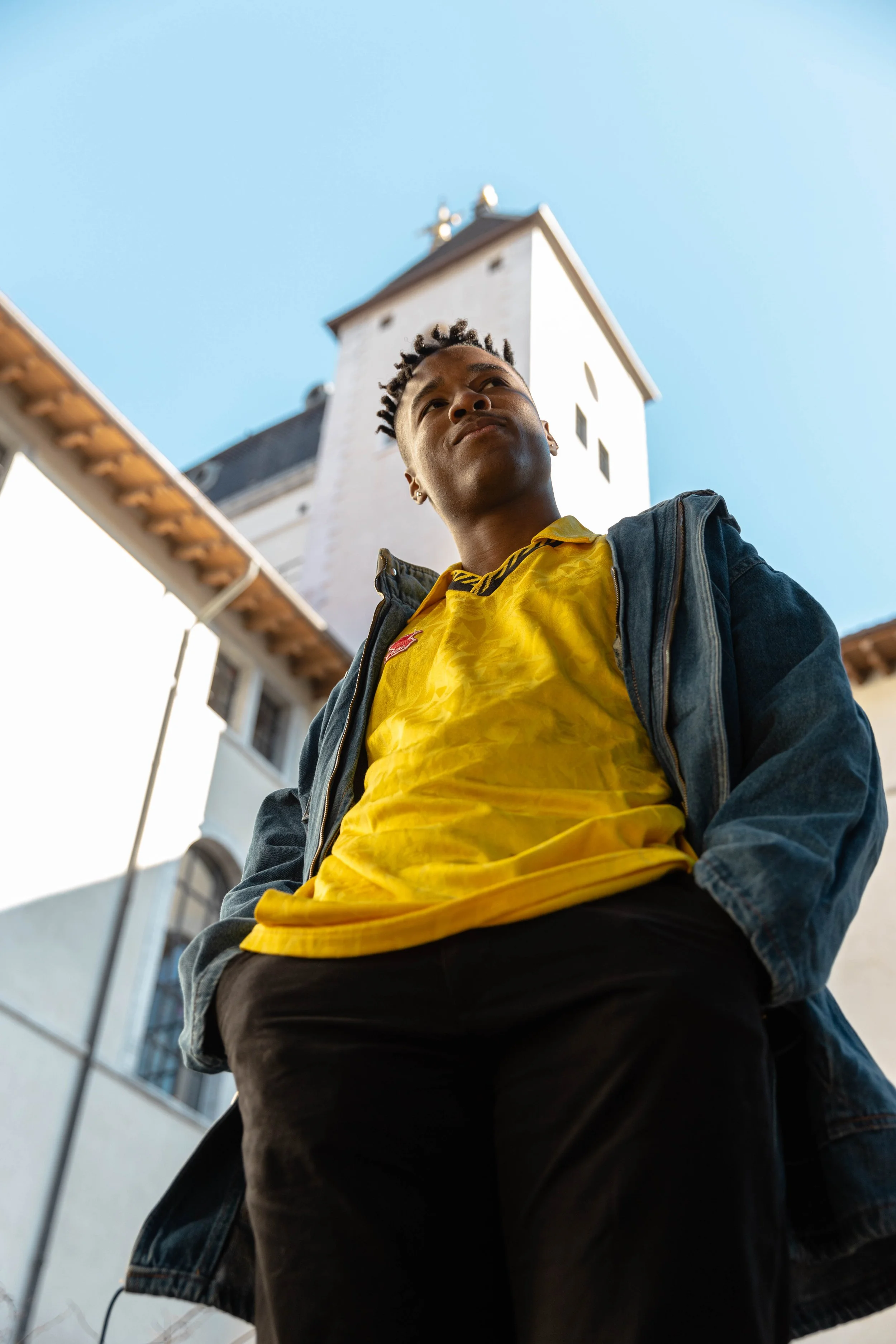 Un jeune homme debout regardant vers le haut, en vêtements décontractés, avec une église blanche en arrière-plan sous un ciel bleu.