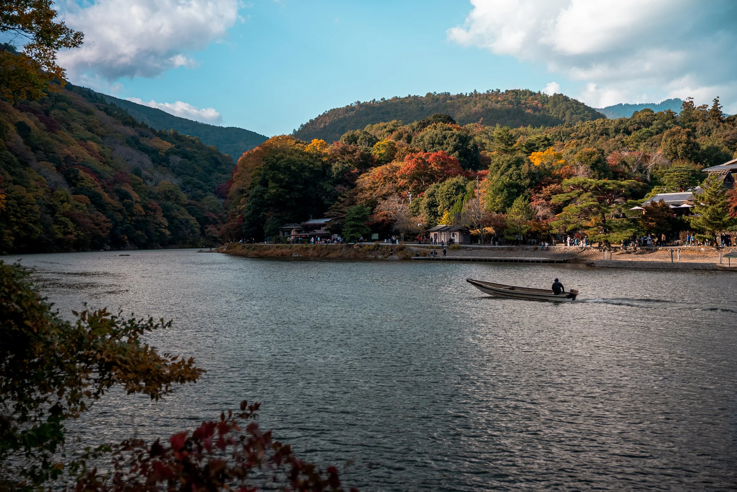 Scène paisible d'une rivière entourée de montagnes et de forêts aux couleurs d'automne, avec un petit bateau naviguant et des bâtiments traditionnels en arrière-plan.