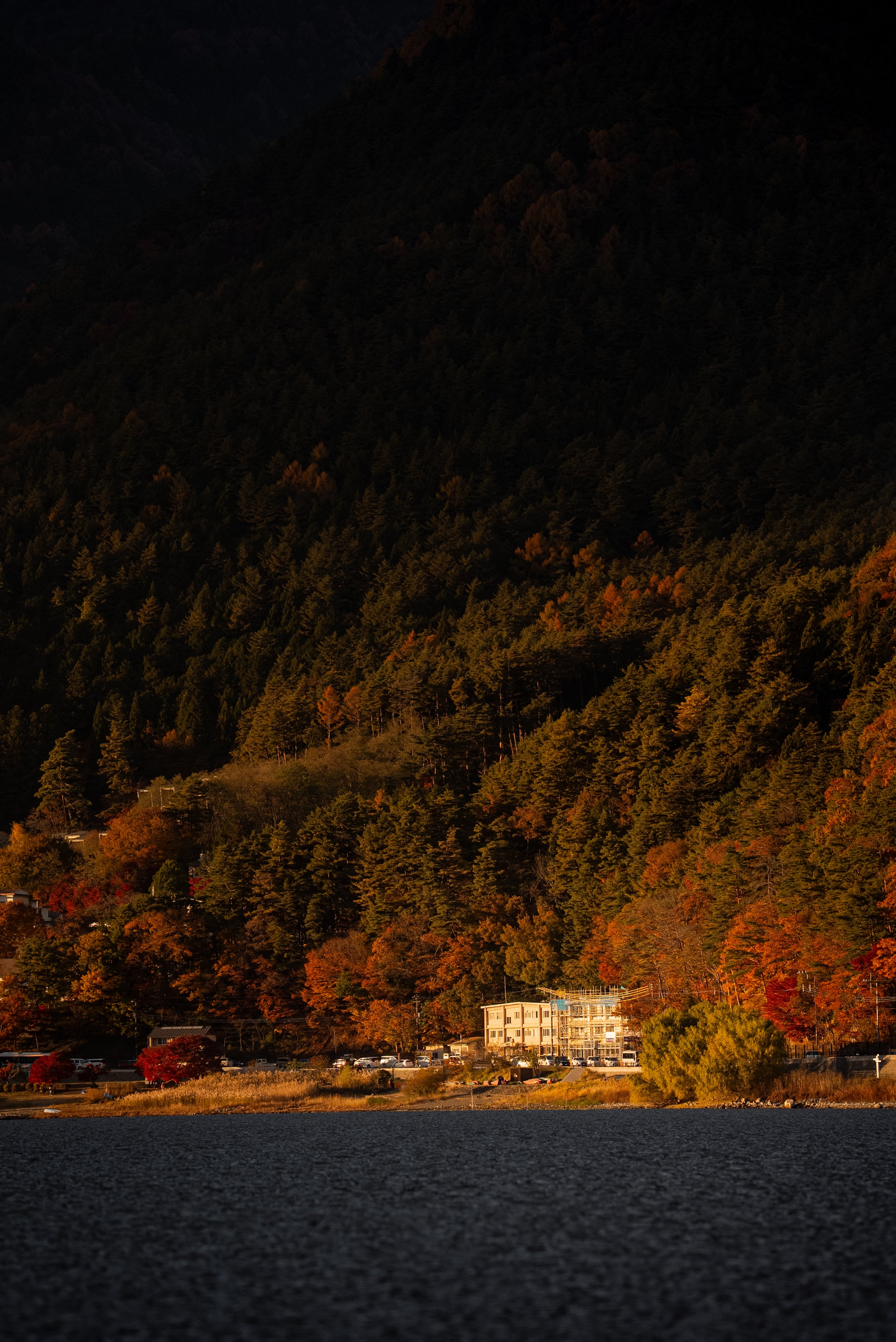 Une scène automnale avec une forêt aux couleurs d'automne, un lac au premier plan, et quelques bâtiments près du rivage, dans un environnement de montagne.