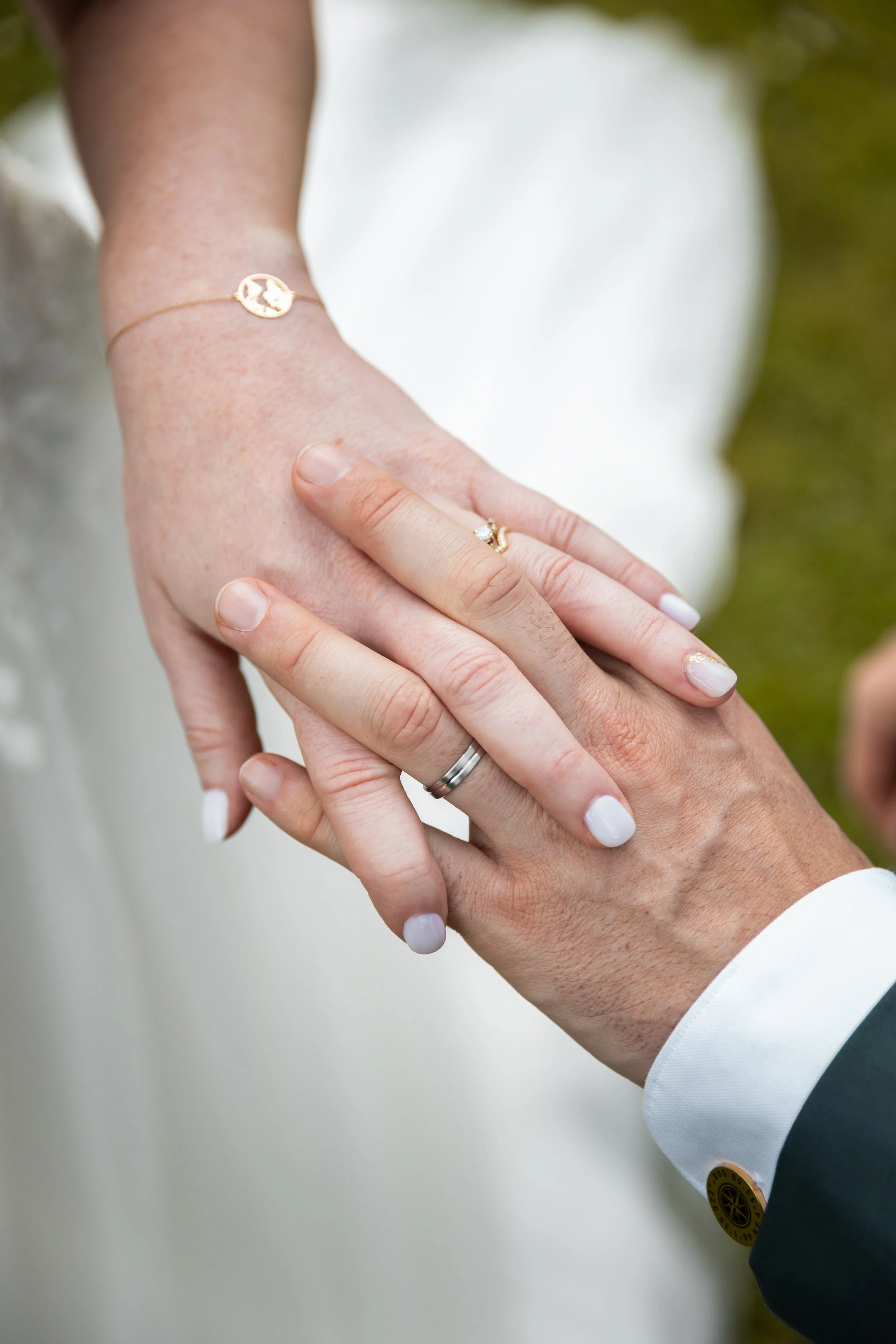 Les mains d'un homme et d'une femme entrelacées, portant des alliances, lors d'un mariage. La femme porte également un bracelet doré avec un pendentif.